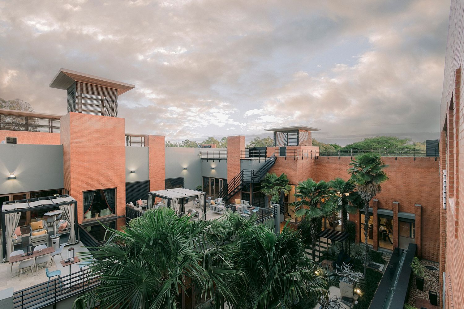 Elevated view of a modern red brick courtyard with outdoor seating, lush palm trees, and open-air walkways under a cloudy dusk sky.