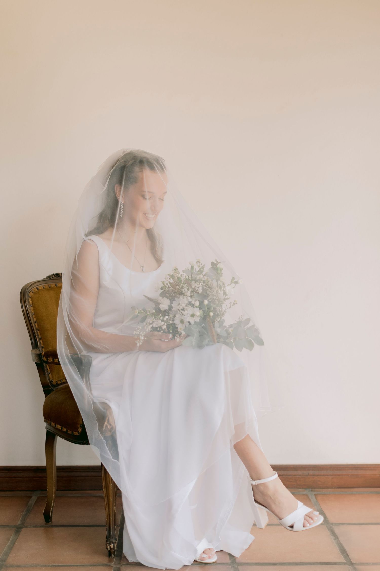 Elegant bride in white gown and veil seated on vintage chair holding delicate floral bouquet.