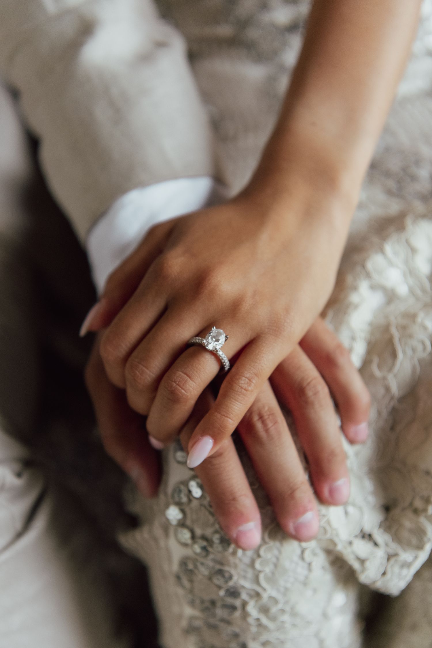 A hand wearing a white engagement ring with a sparkling diamond rests on delicate white lace fabric.