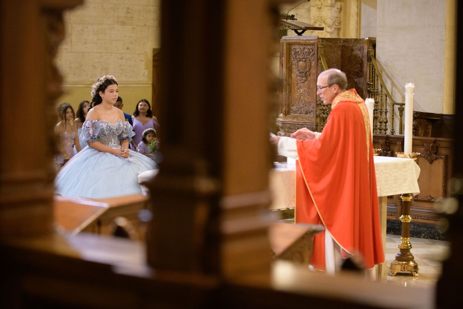A church ceremony in progress with a priest in red robes and participants visible through wooden pews.
