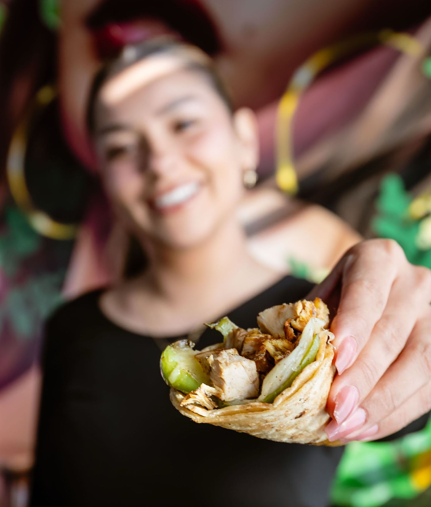 Hand holding a fresh taco filled with meat and vegetables while someone stands in the background.