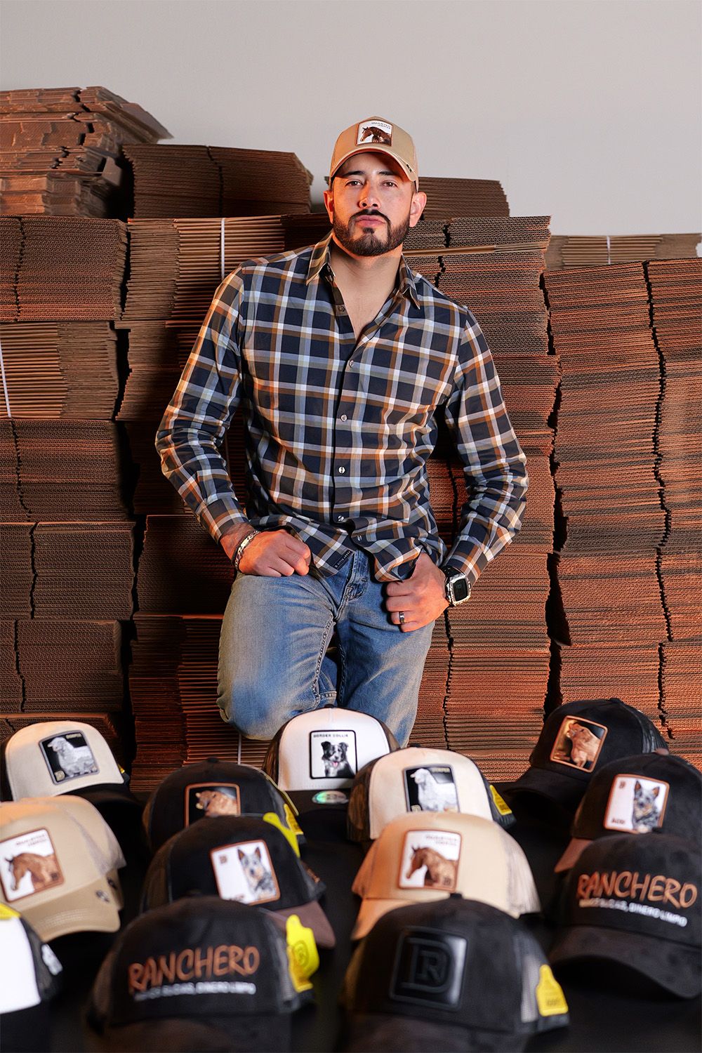 Person in plaid shirt standing against stacked boxes with collection of Panchero branded baseball caps displayed below.