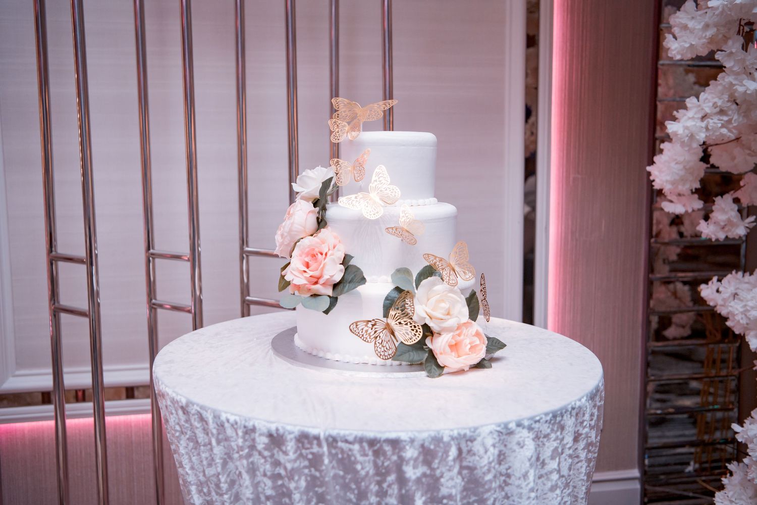 Elegant white wedding cake decorated with fresh pink and white roses sits on lace-covered table with soft pink lighting.