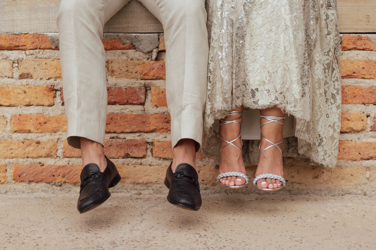 A close-up photo of two pairs of feet in wedding attire against a rustic brick wall.