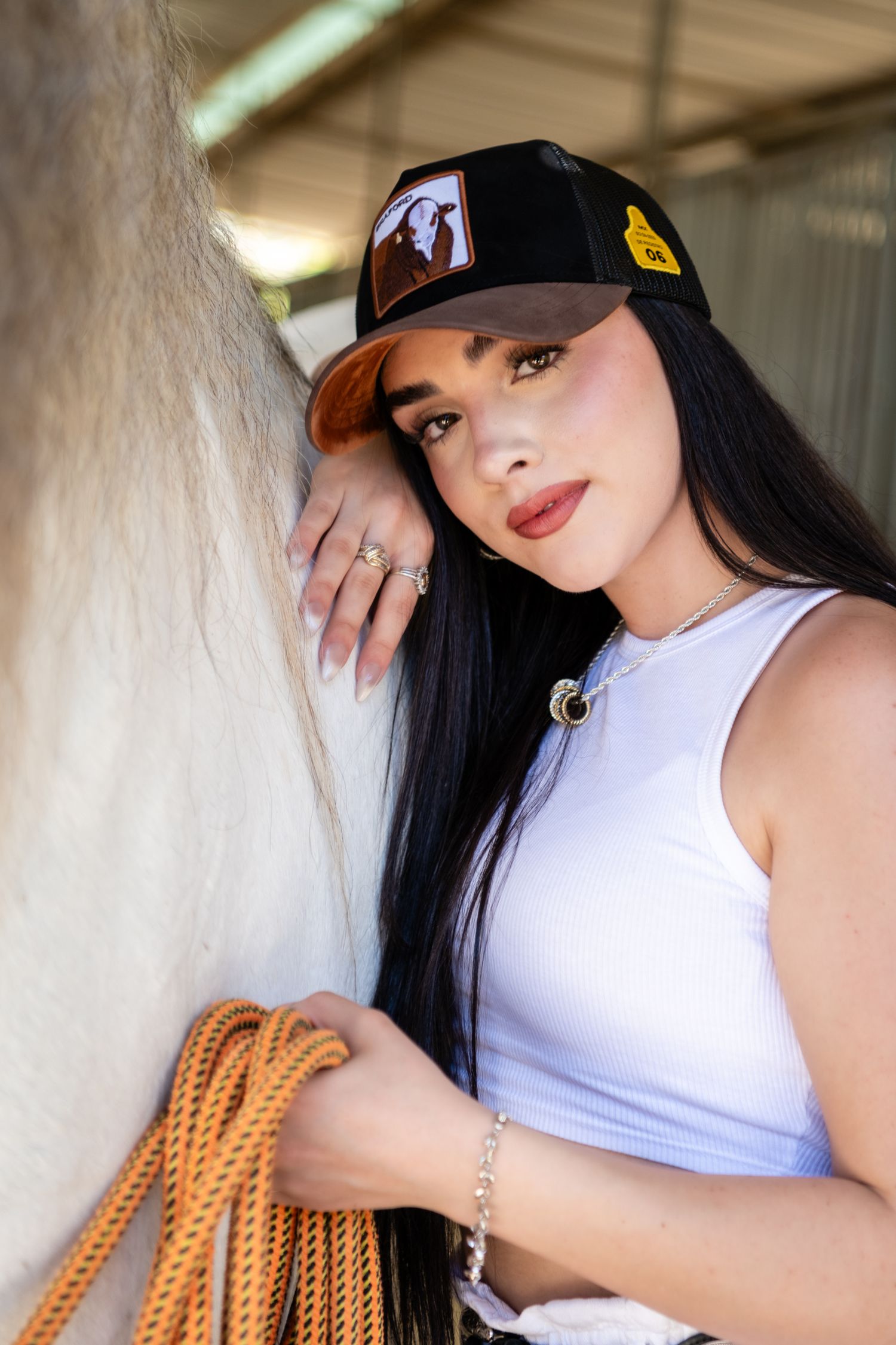 Portrait of a young woman in a white tank top and baseball cap holding orange horse reins at a stable.