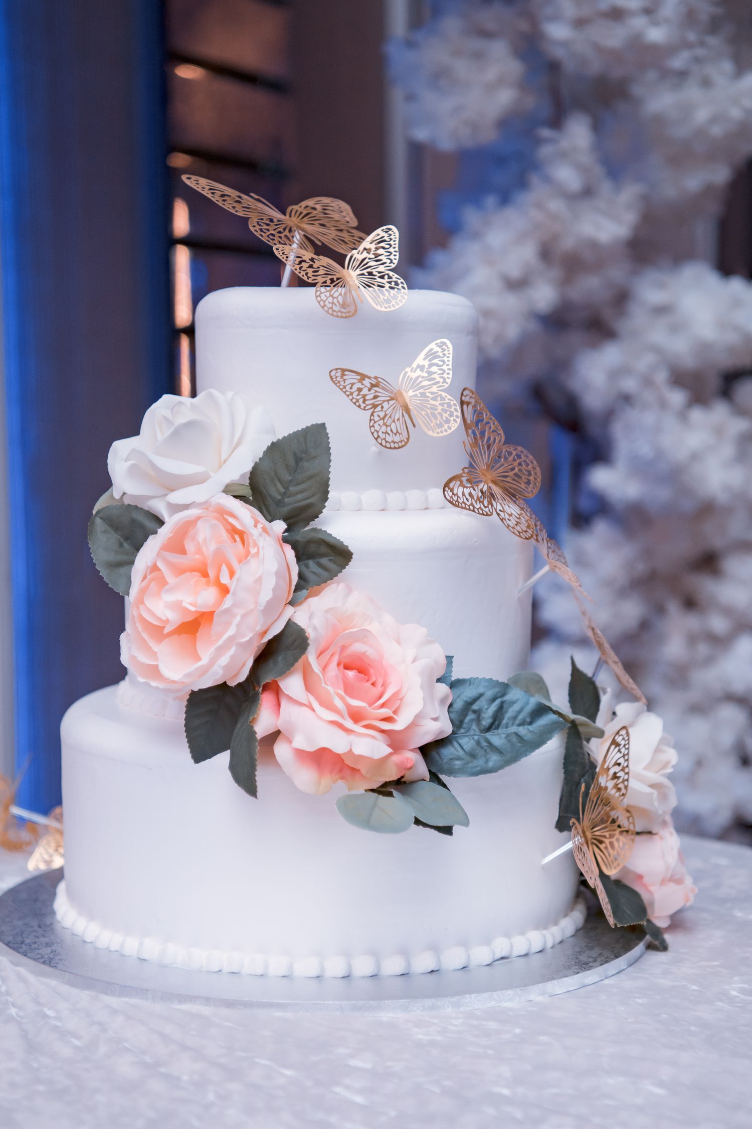 Elegant white tiered wedding cake decorated with pink roses and metallic rose gold butterfly accents.
