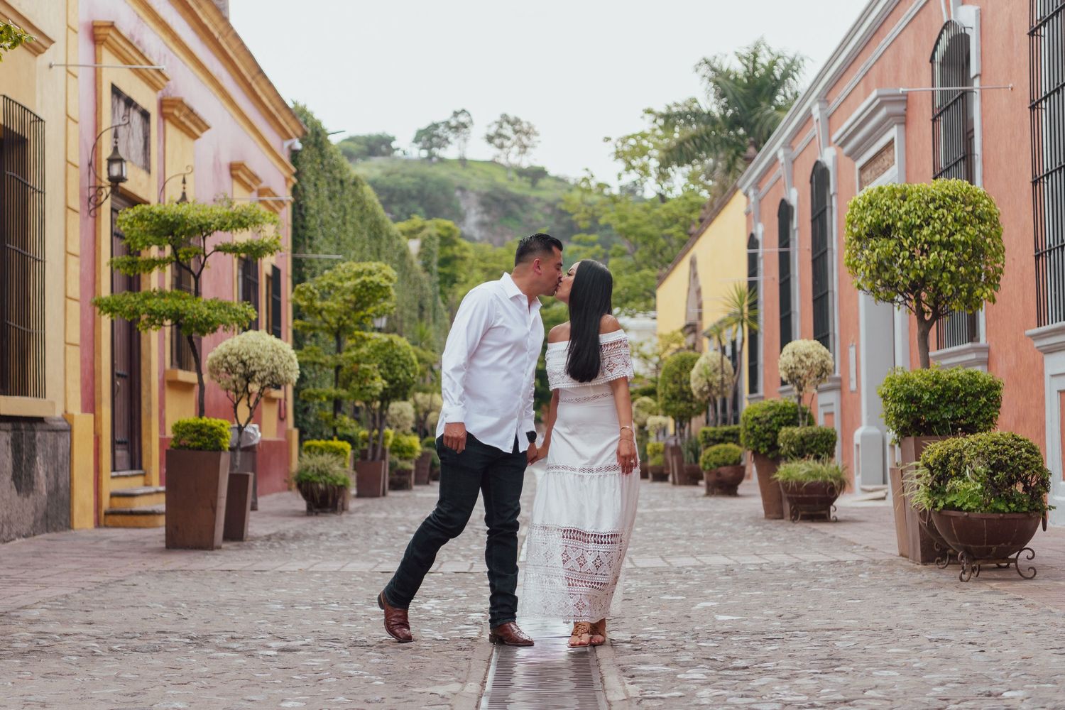 A couple walks down a charming cobblestone street lined with colorful colonial buildings and potted plants.