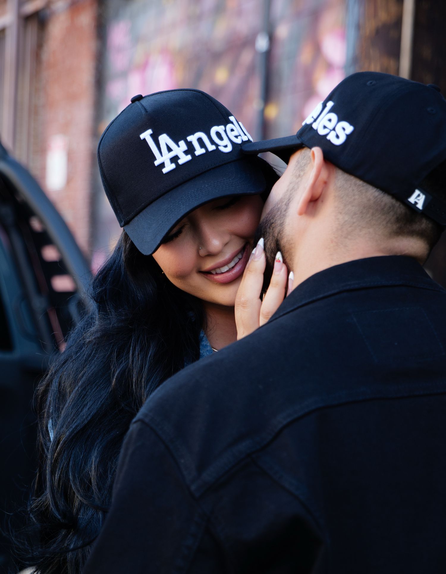 A couple wearing matching LA baseball caps share an intimate moment on a city street.