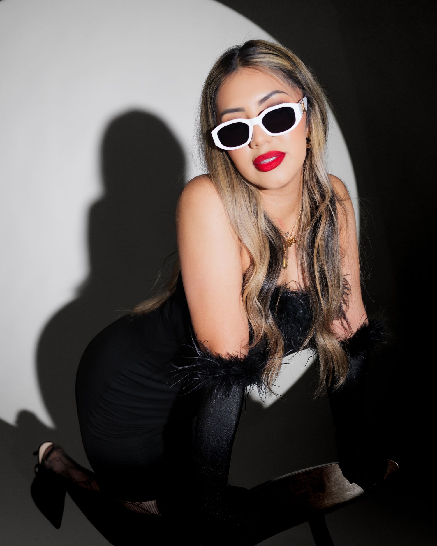 Stylish woman in black dress and white sunglasses poses against white wall with dramatic shadow effect.