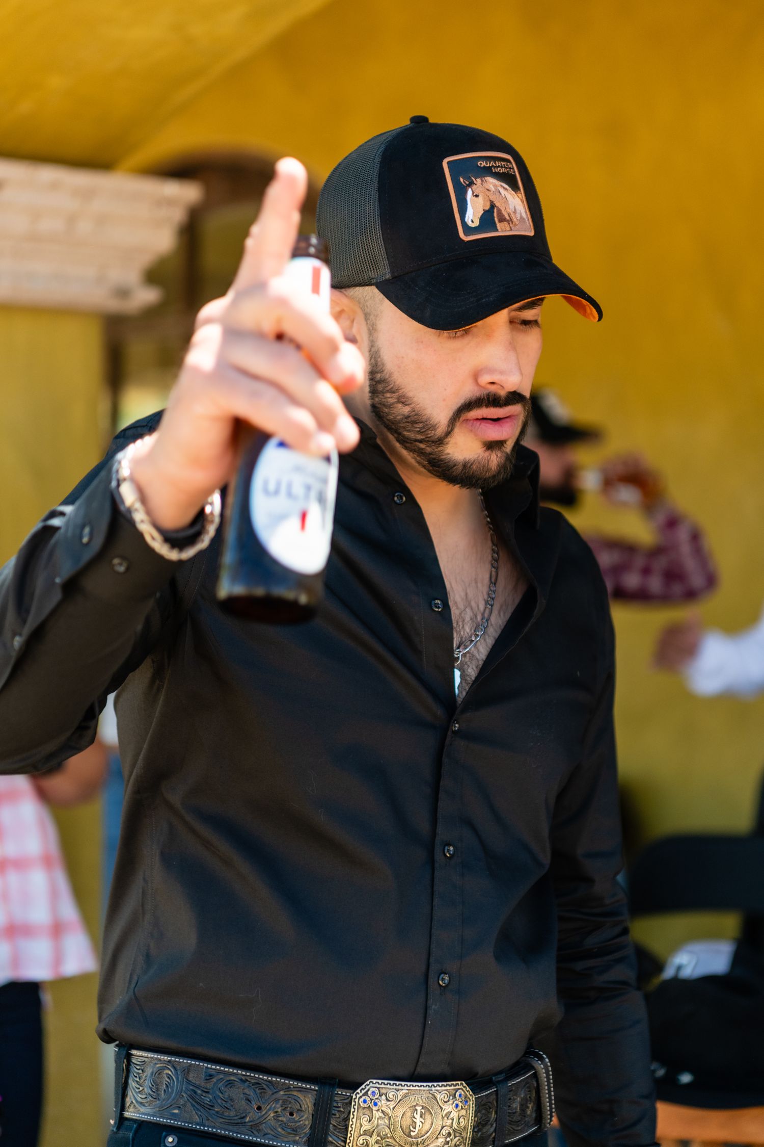 Person wearing black button-up shirt and baseball cap gestures while holding beverage at outdoor event.