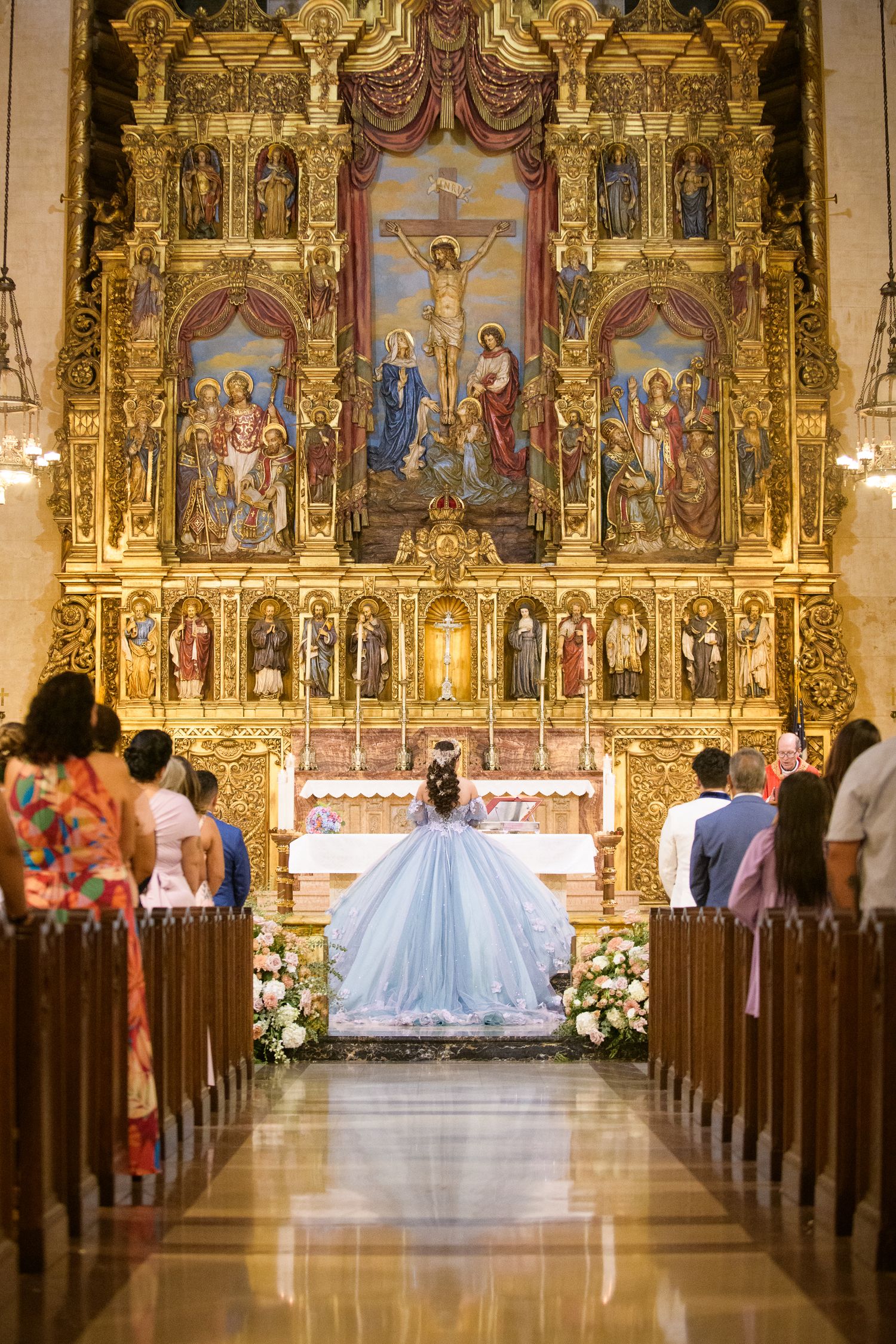 A quinceañera celebration in a Catholic church with an ornate golden altar and religious artwork depicting the crucifixion.