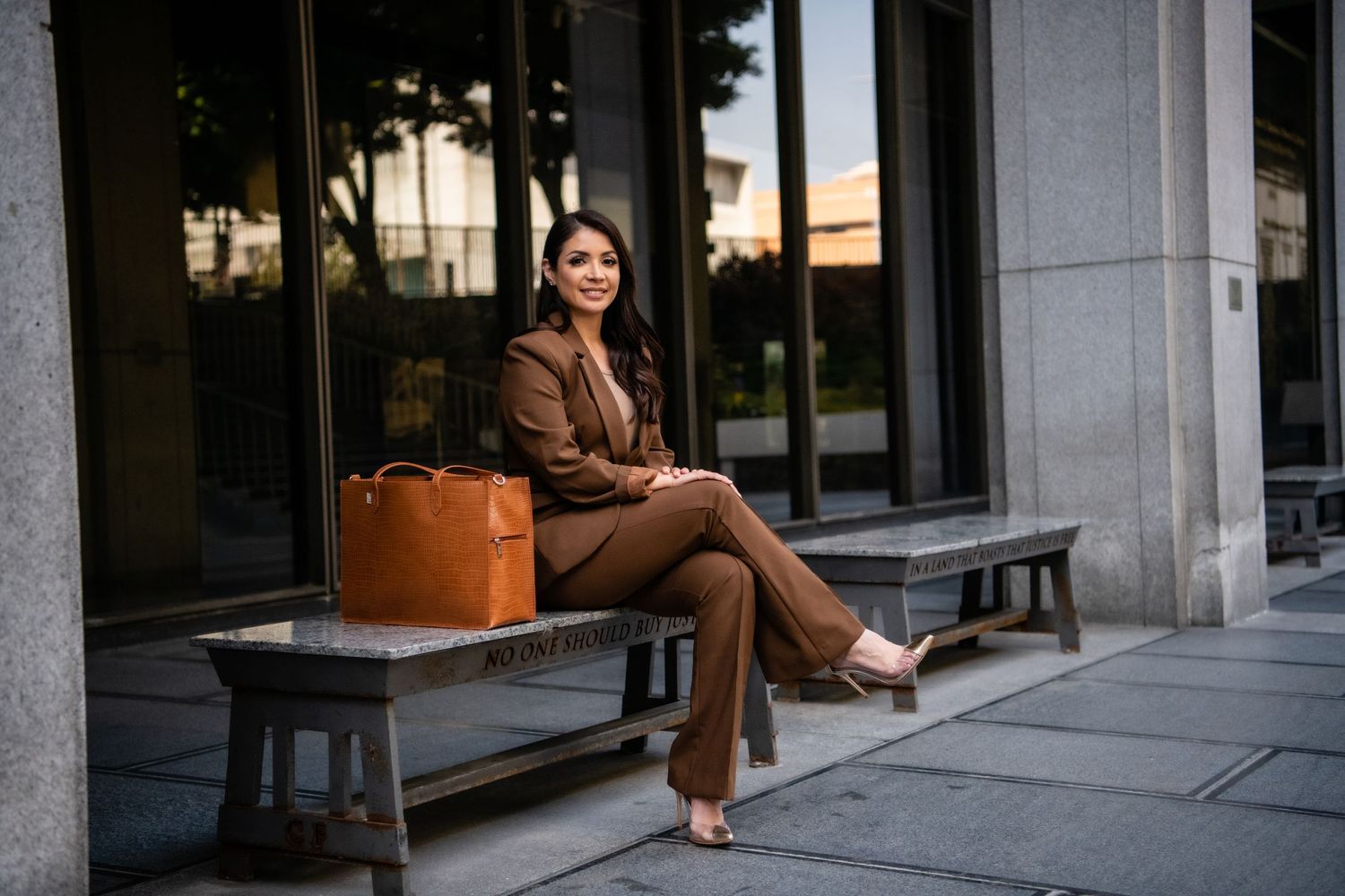 Business woman in brown suit sitting on bench with leather bag outside building.