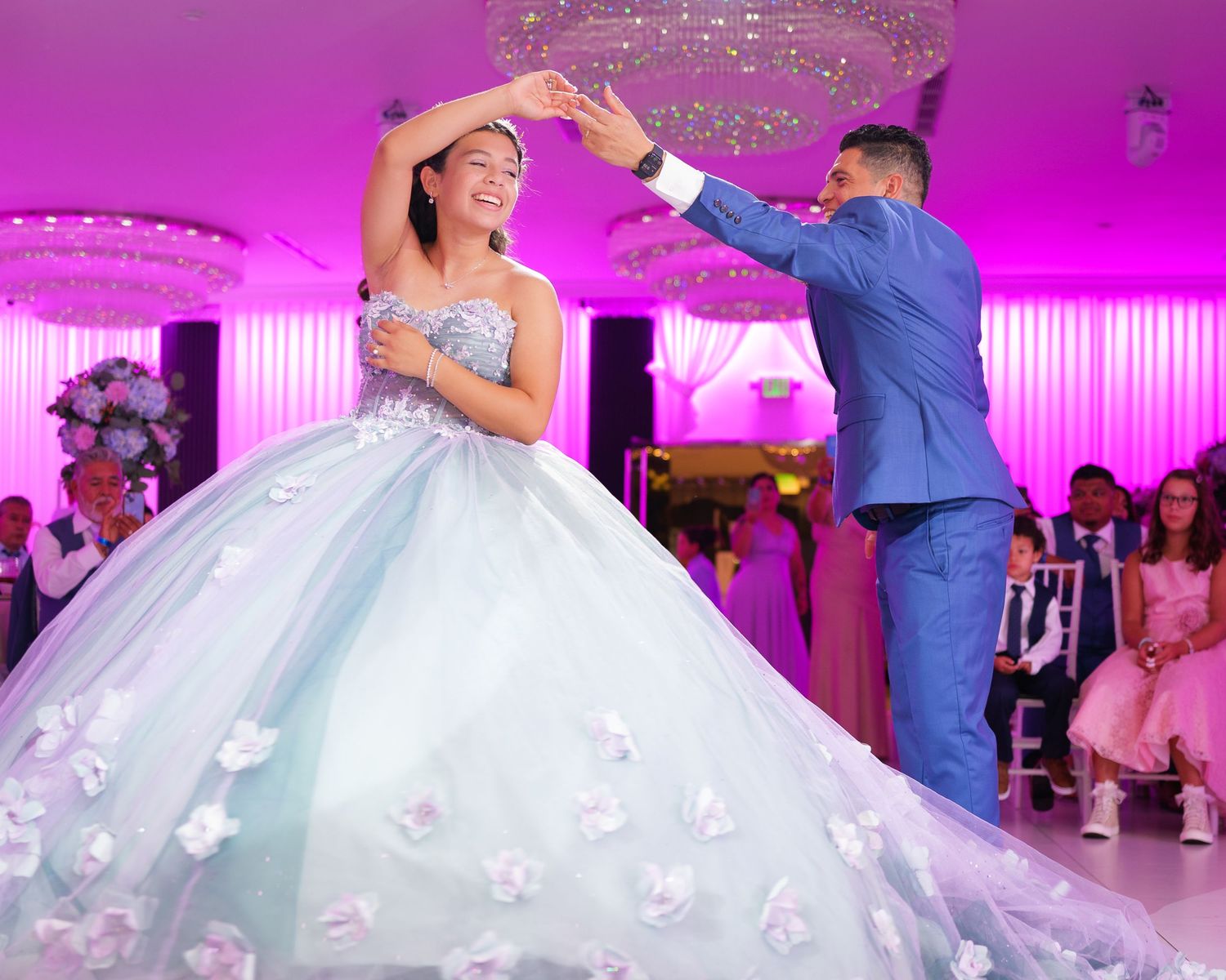 A couple dances at a quinceañera celebration in a ballroom with pink lighting and a flowing light blue ball gown.