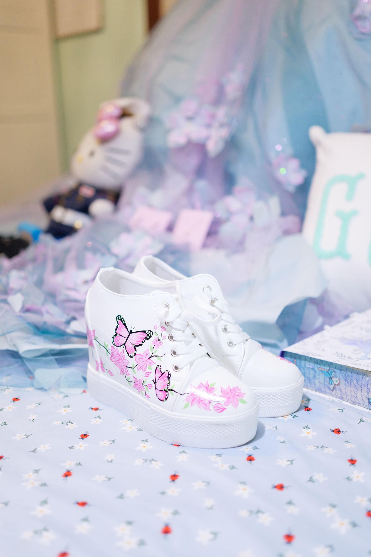 White canvas sneakers with pink butterfly decorations displayed on soft tulle fabric with pastel colors in background.
