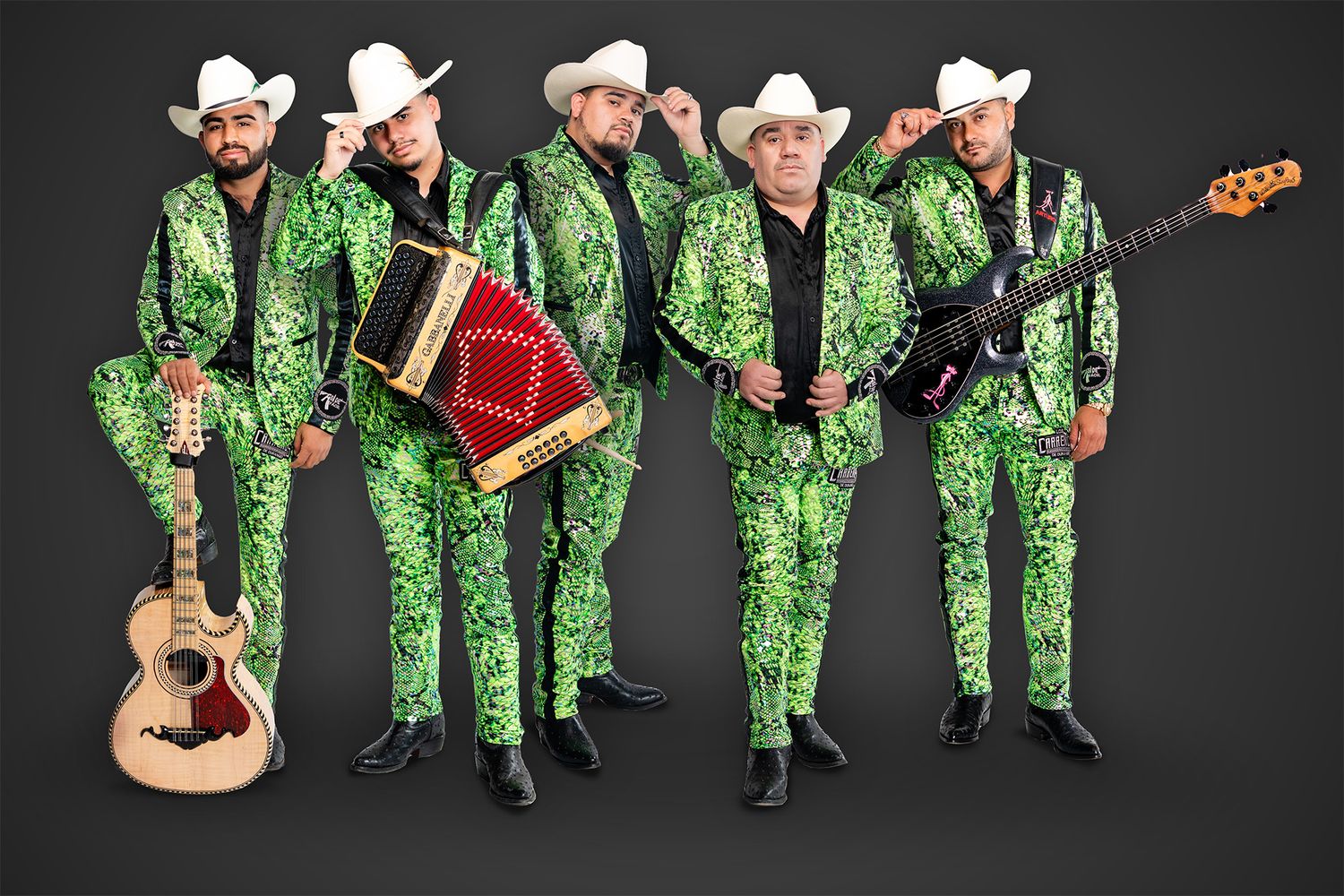 Five musicians in matching green sequined suits and cowboy hats pose with instruments against dark background.