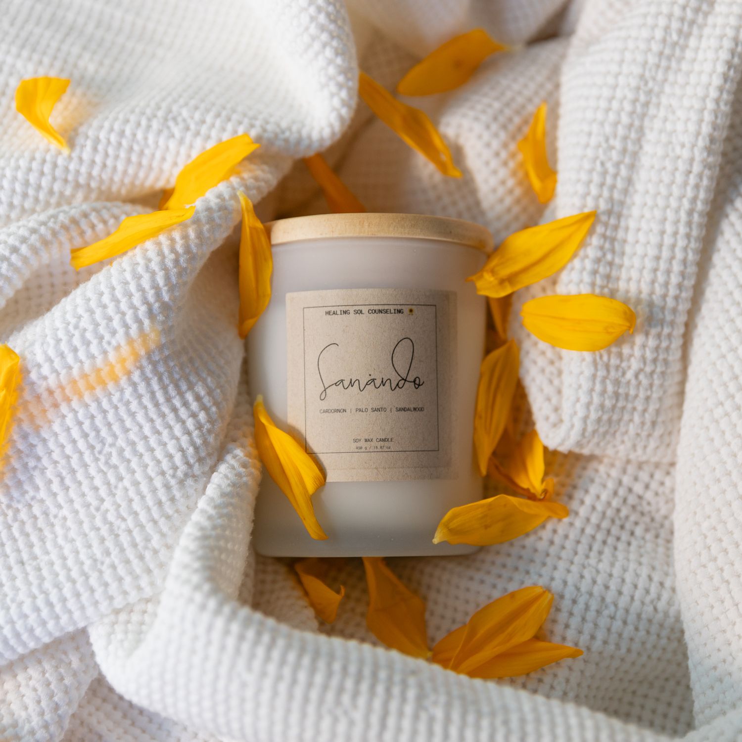 Aesthetic product photography of candle surrounded by yellow flower petals on white knit fabric.