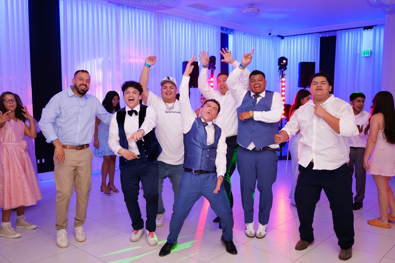 A group of groomsmen and wedding guests dancing energetically on the dance floor with purple lighting at a wedding reception.