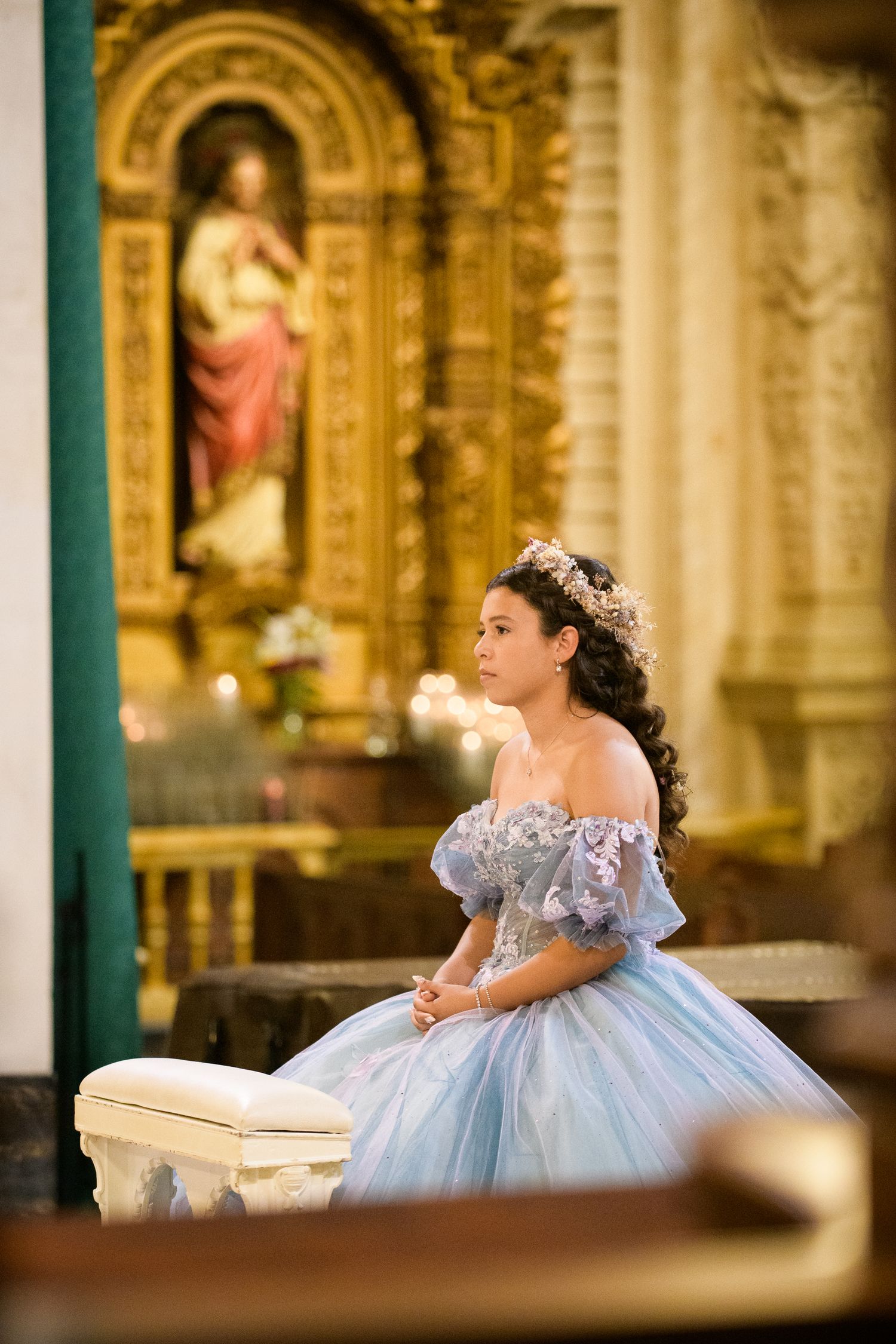 Woman in pale blue ball gown and floral crown poses in an ornate church with golden religious statues in background.
