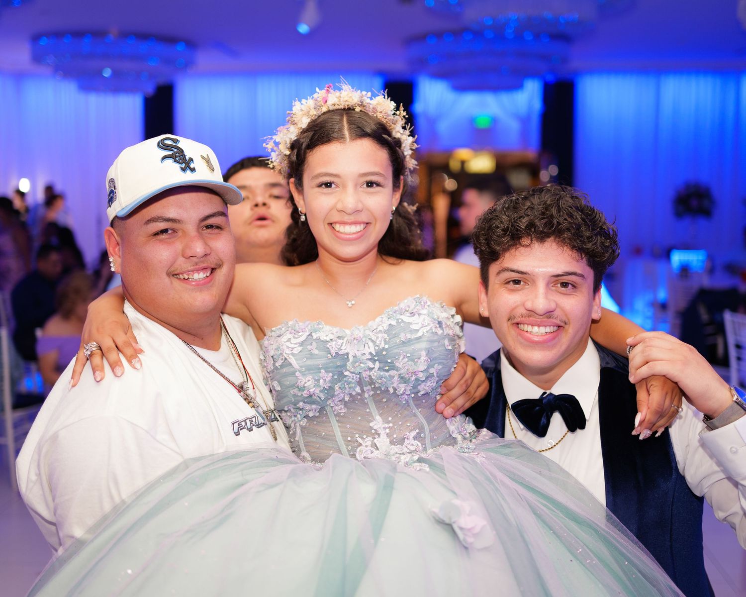 A quinceañera celebration in a ballroom with blue lighting features three people posing together for a photo.
