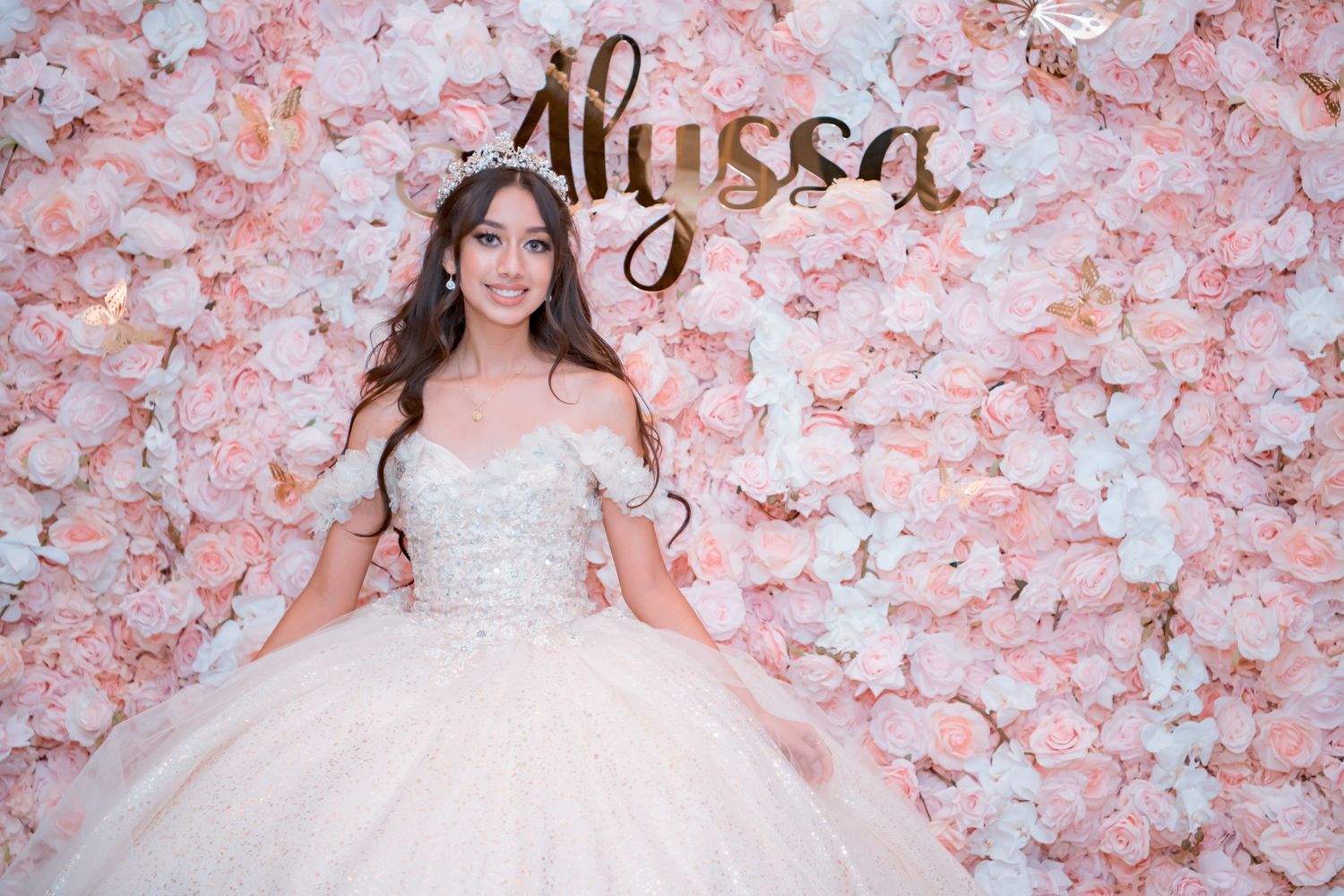 A beautiful bridal gown with off-shoulder details and full tulle skirt against a pink floral wall backdrop.