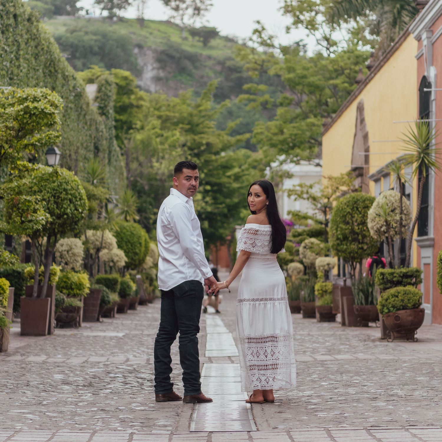 A couple stands holding hands on a cobblestone path lined with decorative potted plants and greenery in a garden setting.