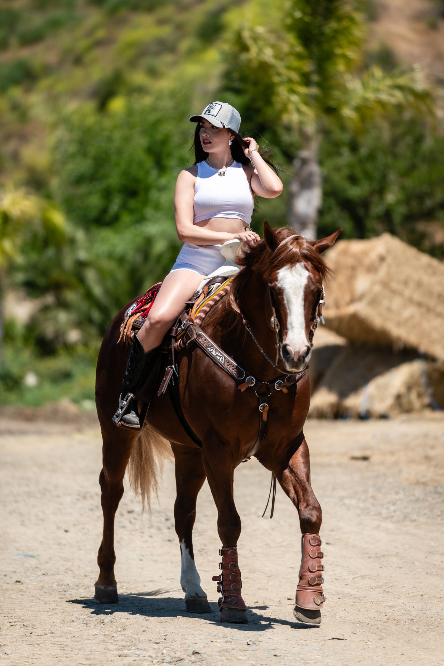 A rider in athletic wear and baseball cap sits atop a brown horse with white markings on a sunny desert trail.