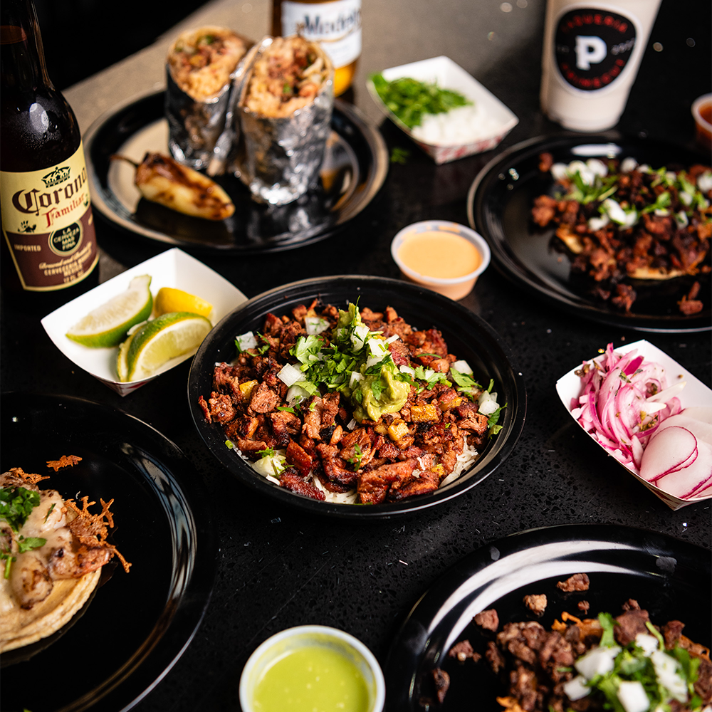 Mexican street tacos and appetizers with tortilla chips, fresh limes, and dipping sauces arranged on black plates.