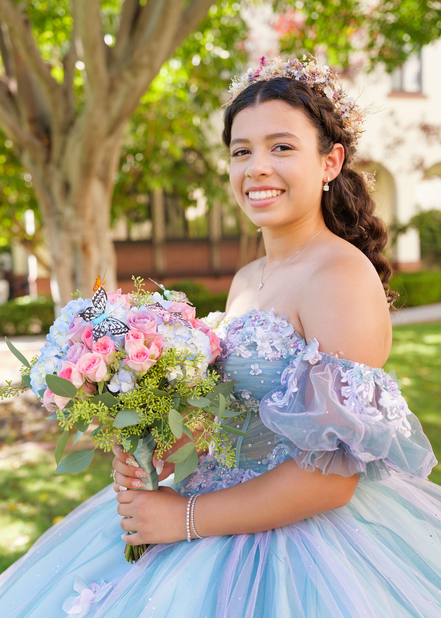 A quinceañera in a powder blue ball gown with off-shoulder details holds a pink and white floral bouquet in outdoor setting.