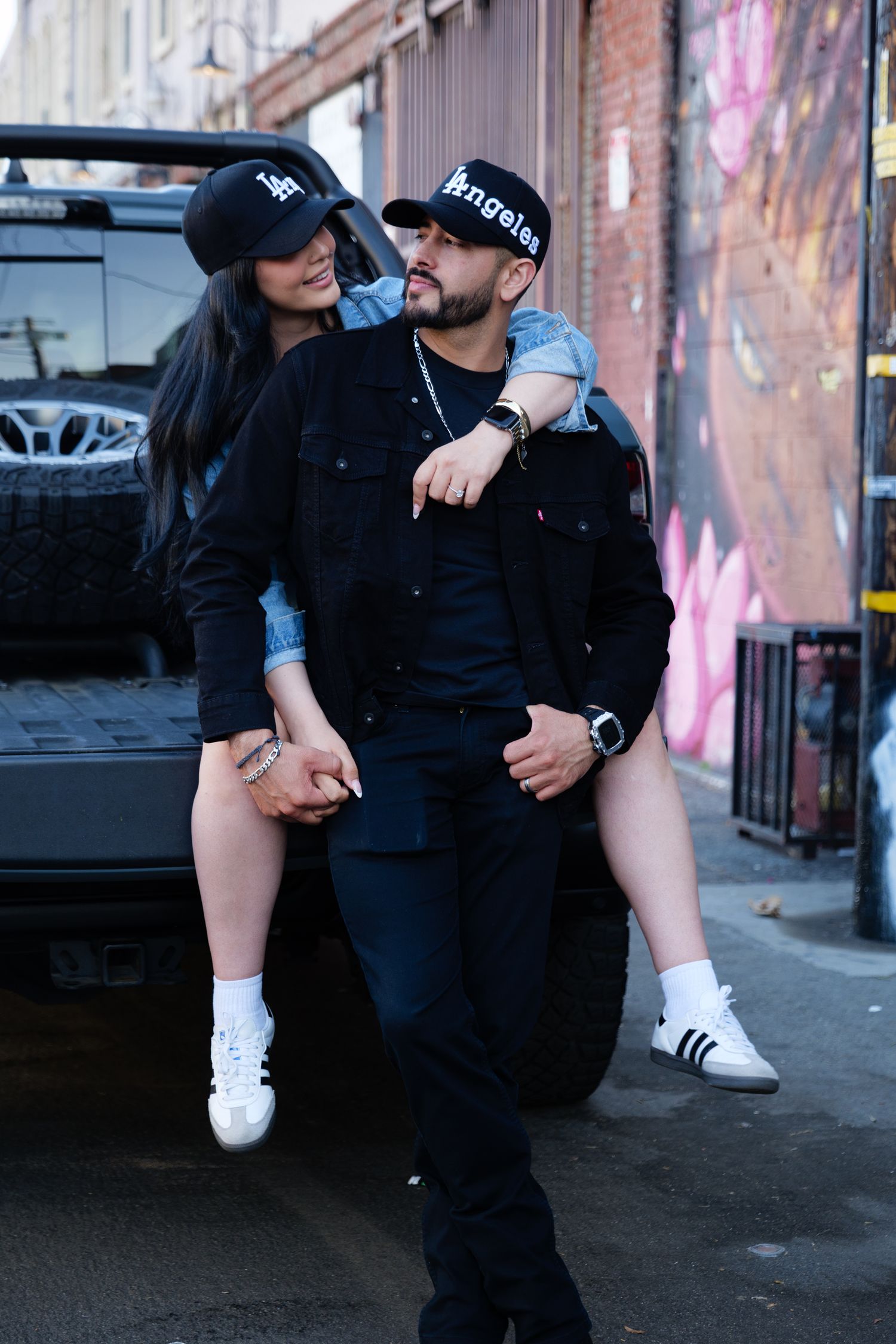A casual couple shares a playful moment while sitting on a pickup truck near a brick wall with street art.