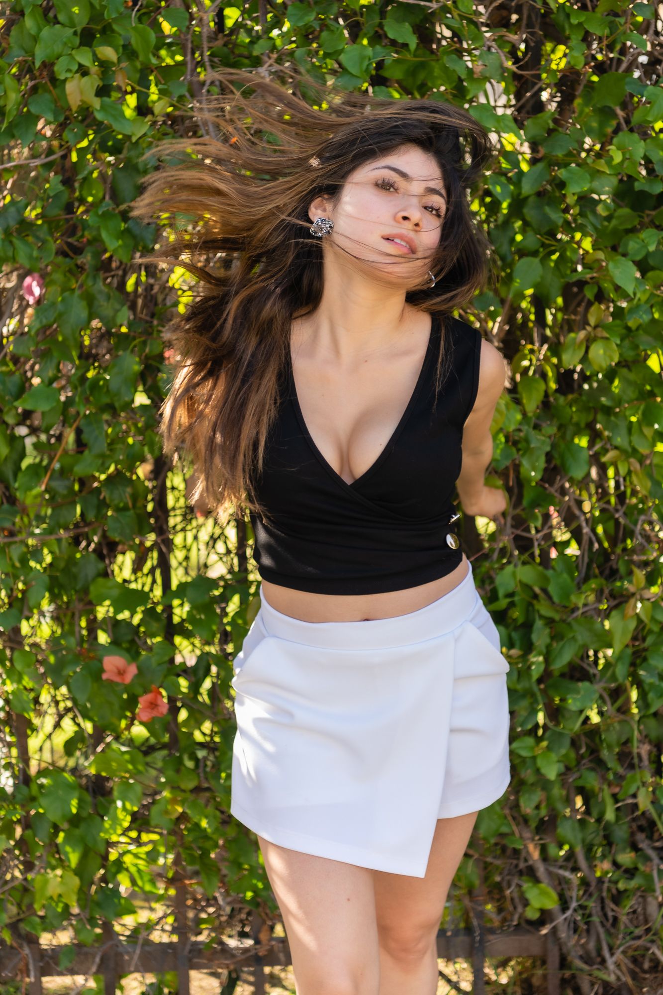Fashion portrait of woman in black top and white skirt with hair blowing in breeze against foliage.