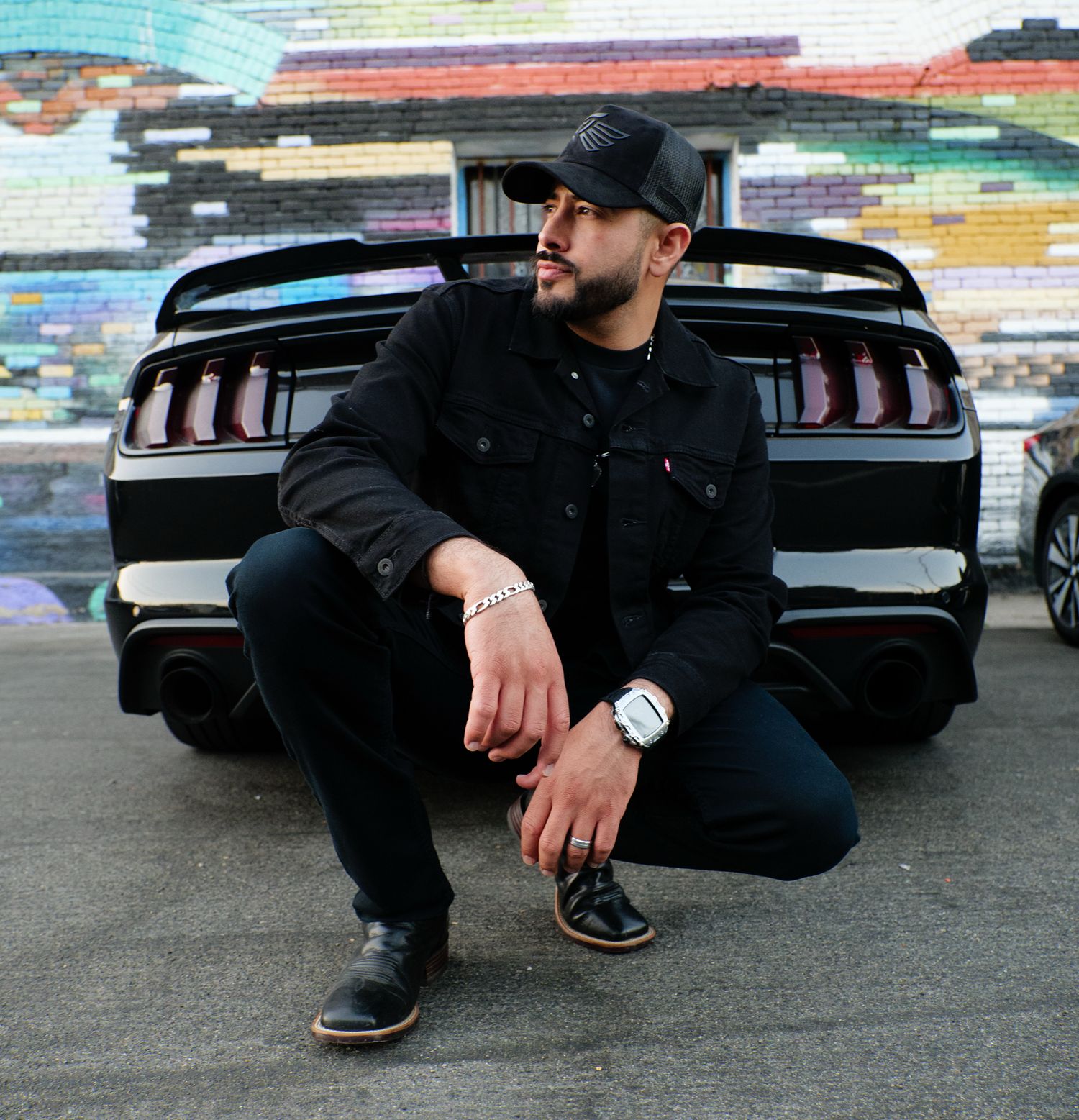Someone crouches in front of a black Ford Mustang with a colorful wall mural in the background.