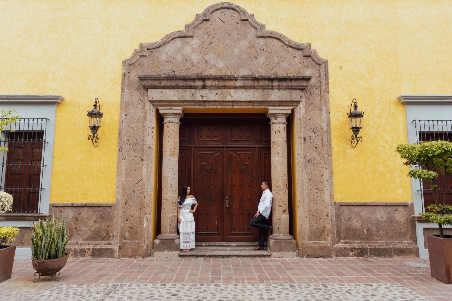 Two figures stand at the entrance of a historic colonial building with a yellow wall and ornate stone doorframe in Mexico.