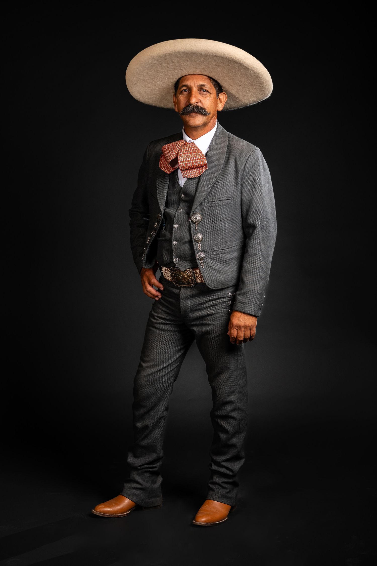 Traditional Mexican charro poses in gray suit and wide-brimmed sombrero against black background.