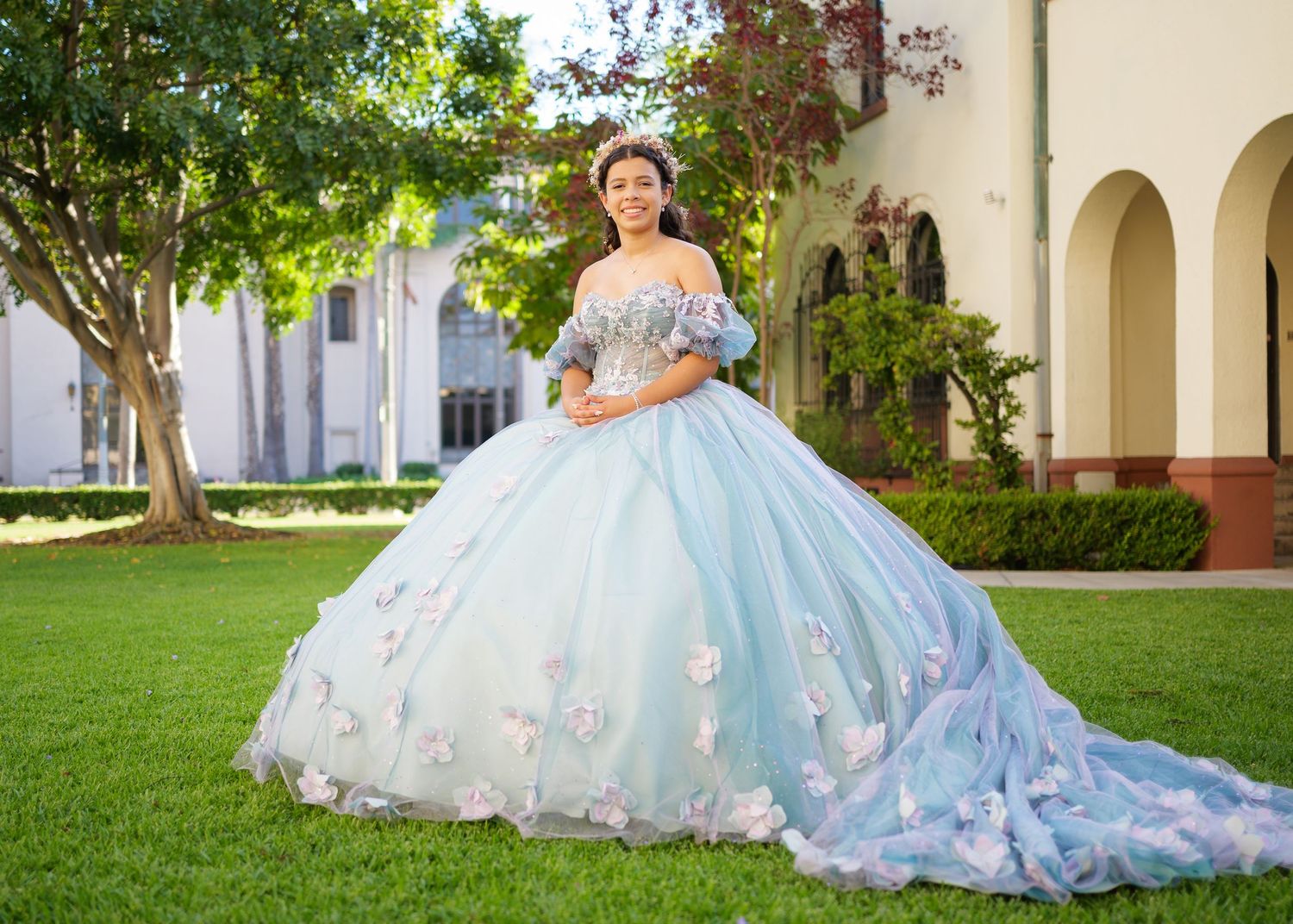 A light blue quinceañera ballgown with floral details and a flowing train displayed on a manicured lawn near arches.