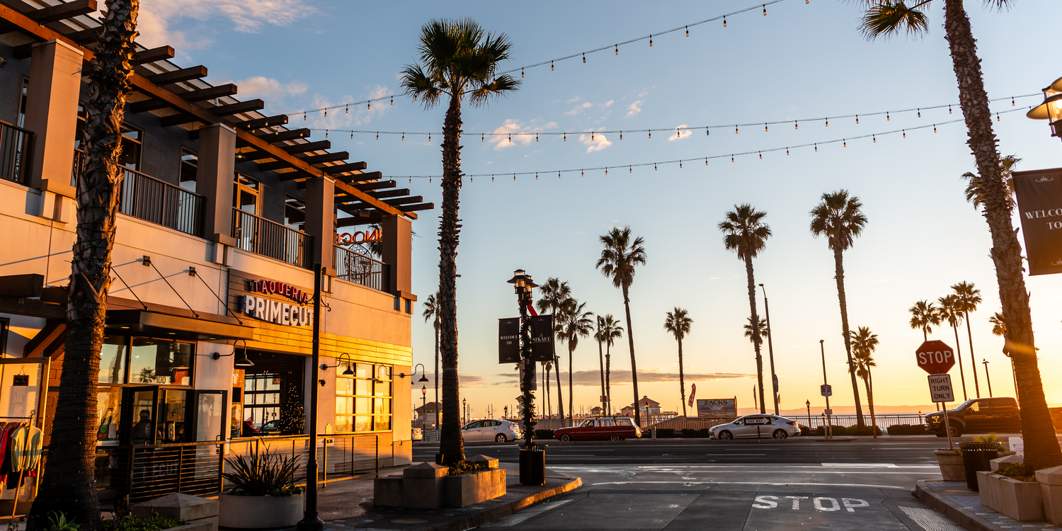 Sunset over beach town promenade with palm trees and string lights illuminating shops and restaurants.
