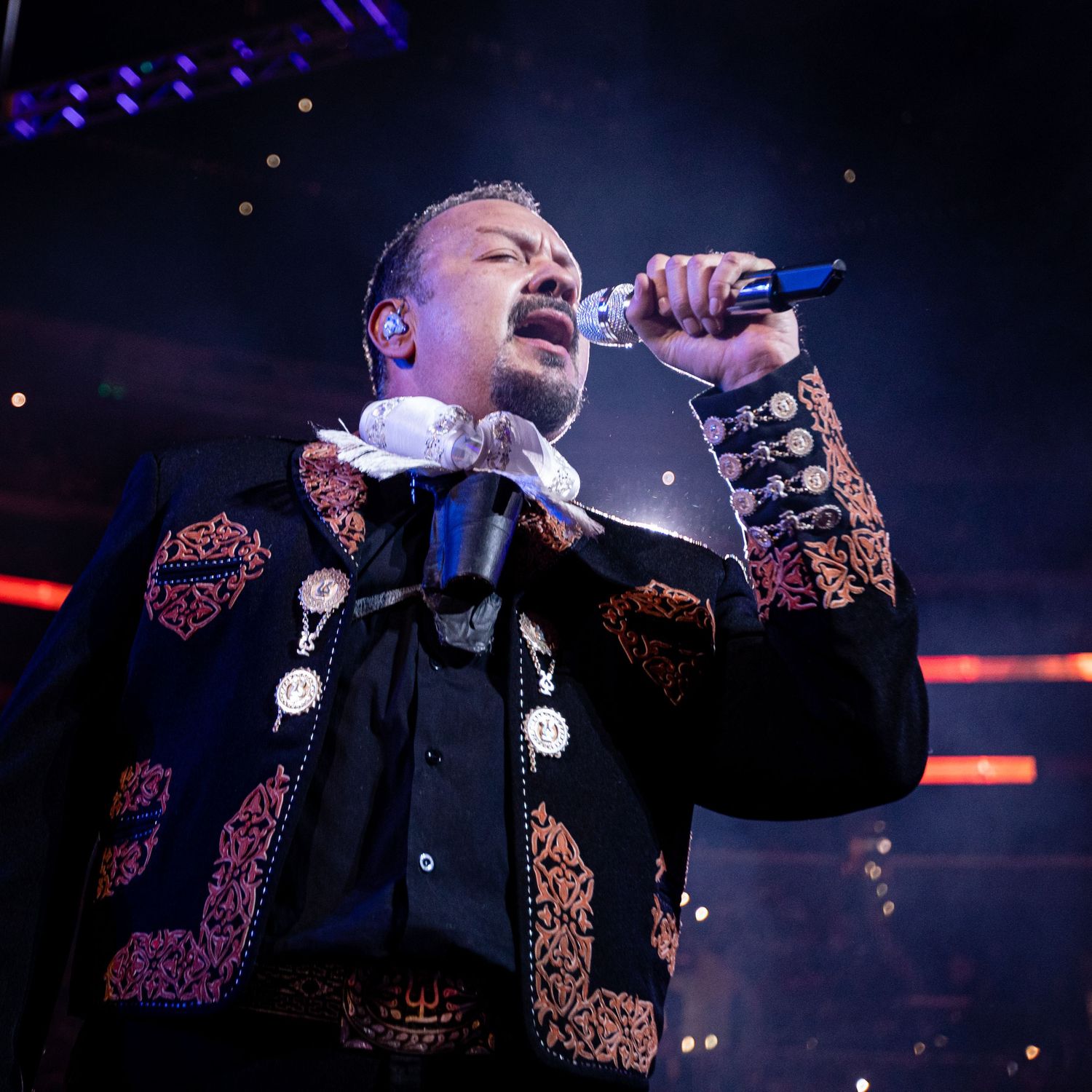A performer in an ornate mariachi-style jacket sings passionately on stage with dramatic lighting.