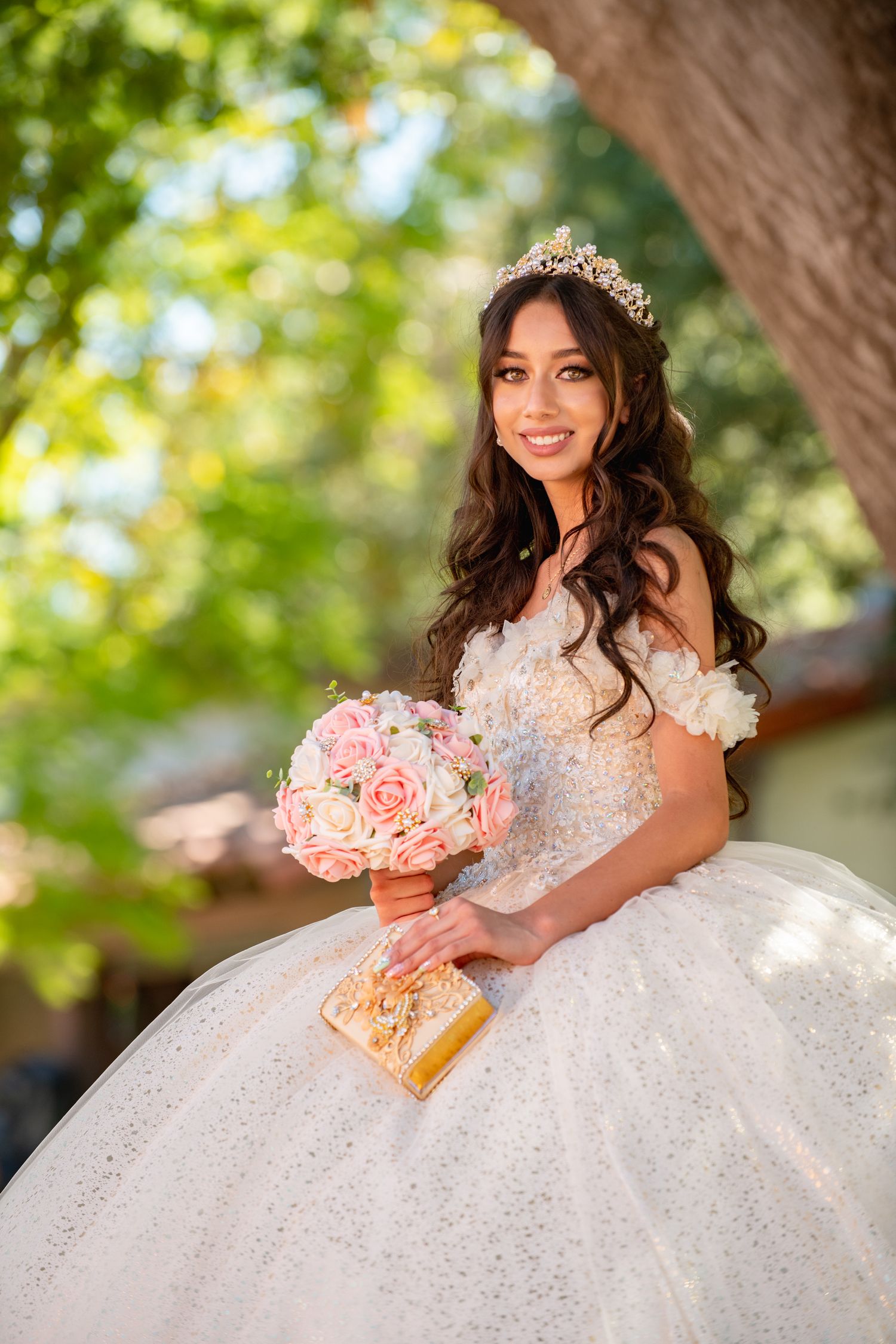 A bride in a sparkly white ball gown and tiara holds a pink rose bouquet while posing outdoors in natural lighting.