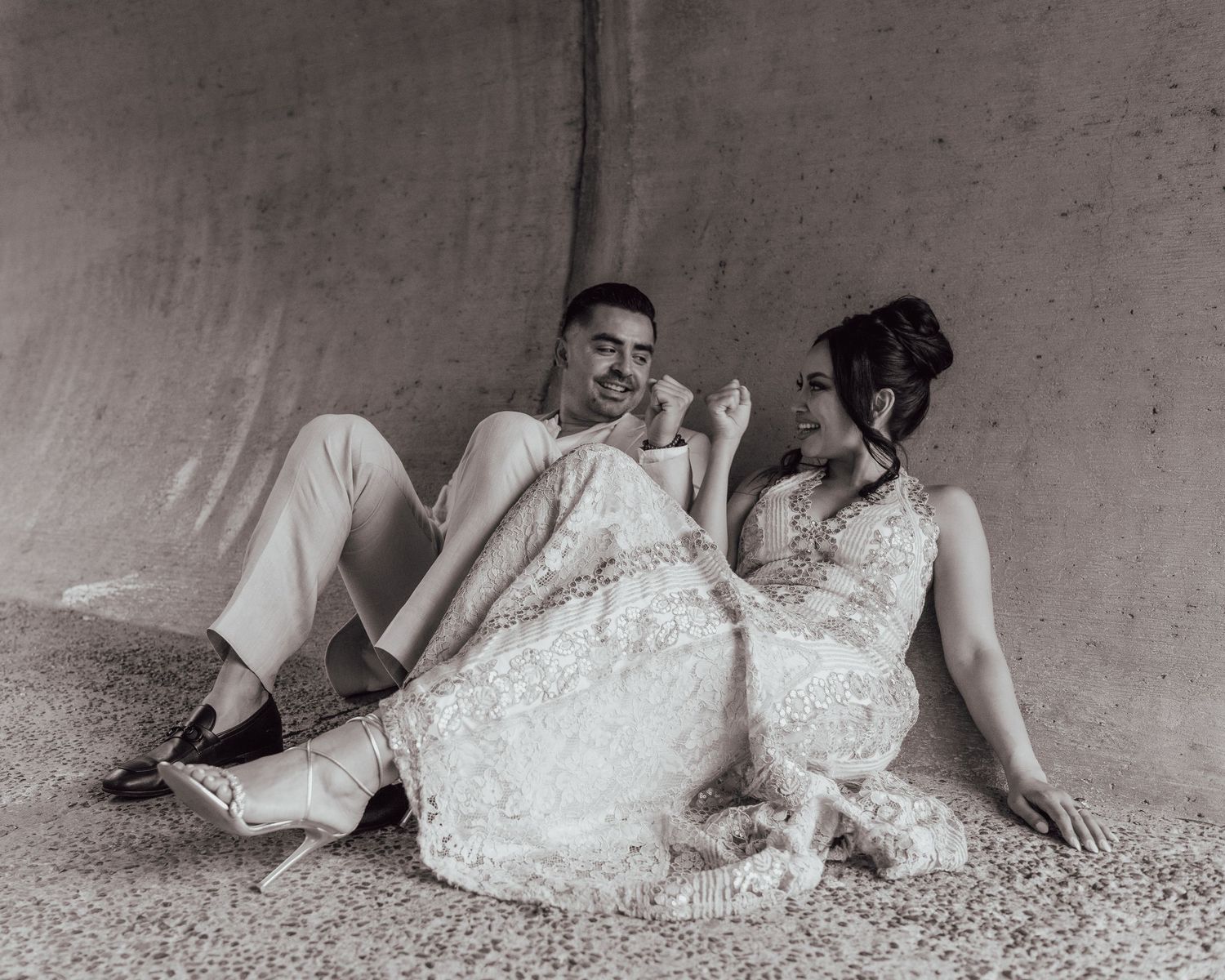 A black and white photo of a couple in elegant white lace outfits sitting on the floor leaning back to back.