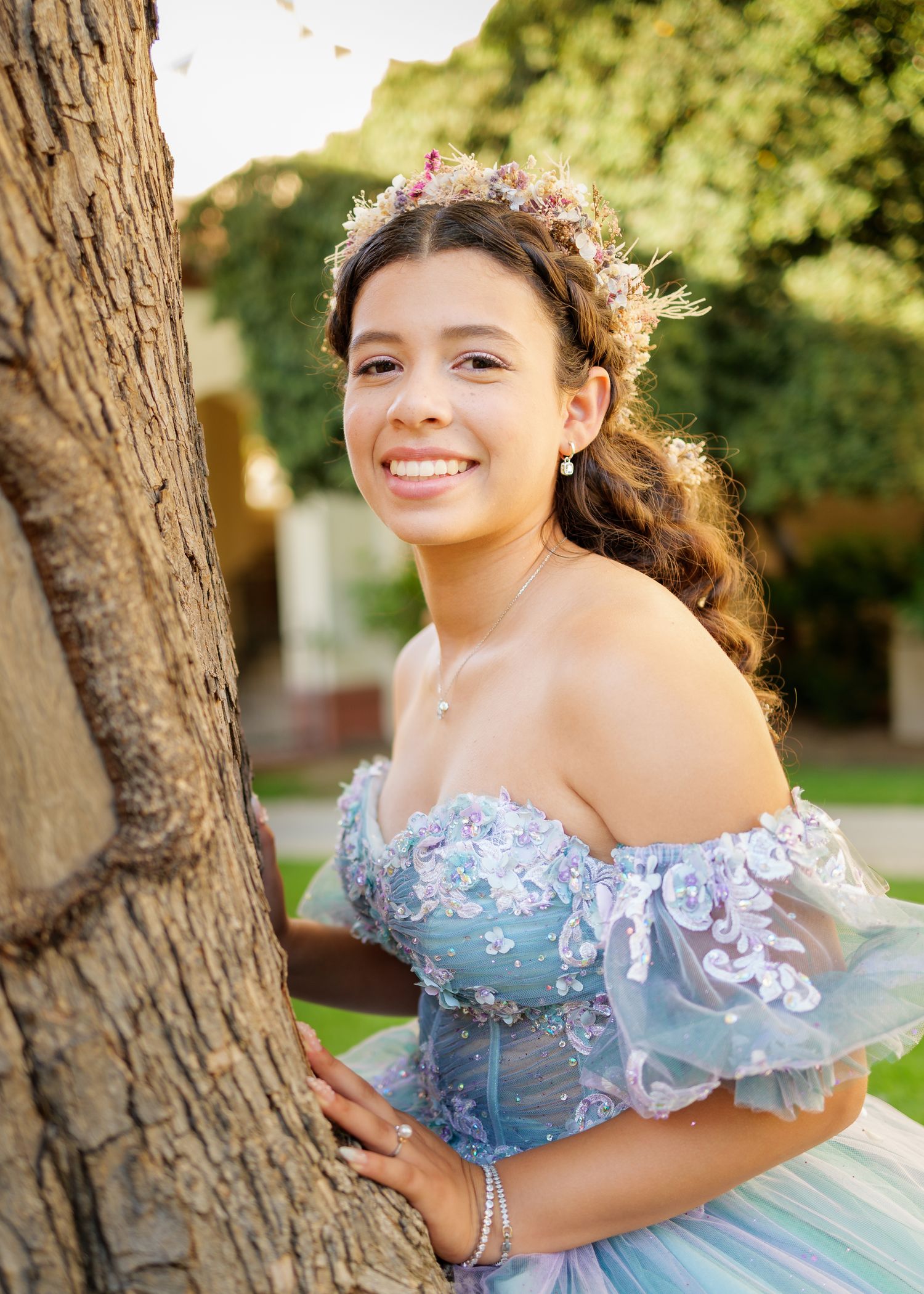 A young woman in a pale blue off-shoulder quinceanera dress stands by a tree trunk in an outdoor garden setting.