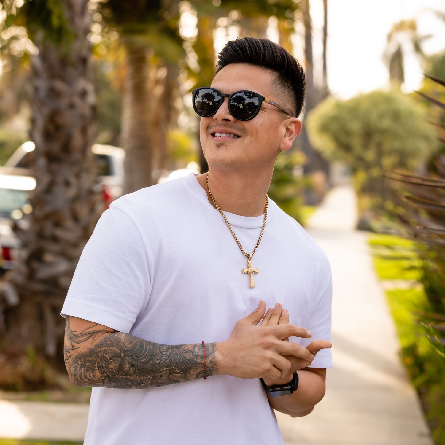 Man in white t-shirt smiling outdoors wearing sunglasses and gold cross necklace.