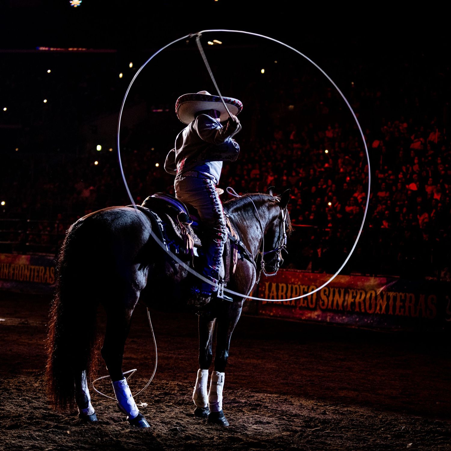 Cowboy performing rope tricks on horseback in dark rodeo arena.
