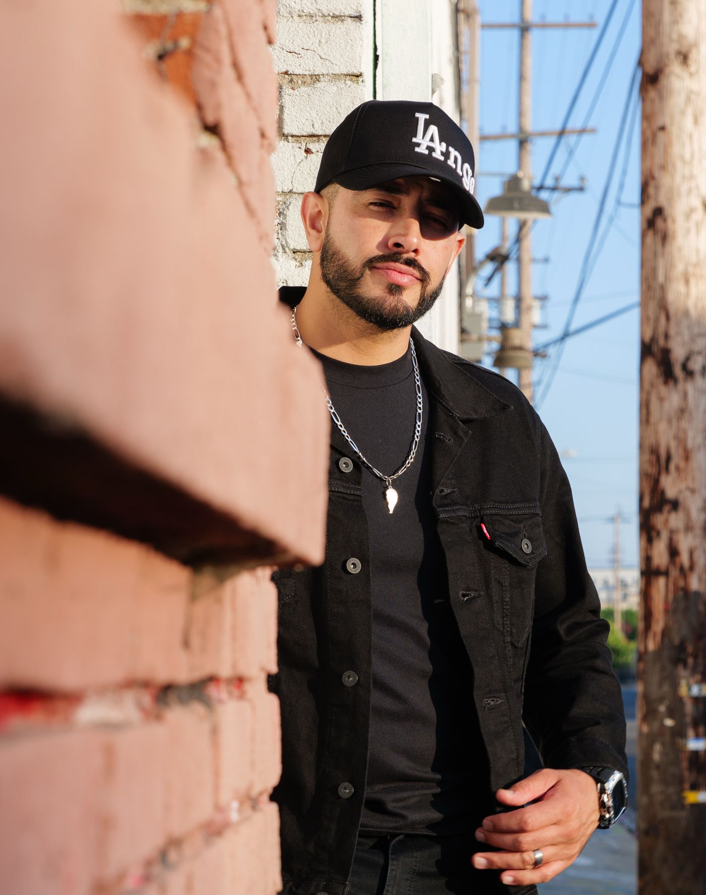 Person in black denim jacket and LA baseball cap poses against brick wall with utility poles in background.