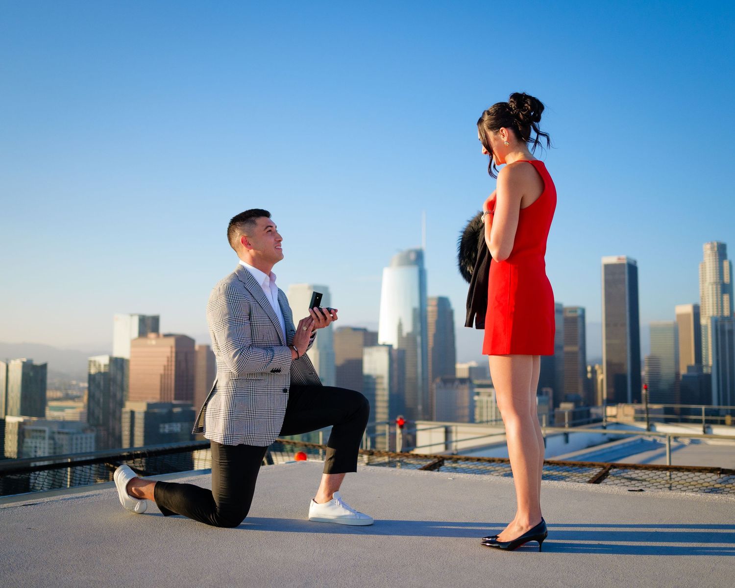 A romantic rooftop proposal in front of a stunning city skyline at sunset with a figure in red dress and grey blazer.