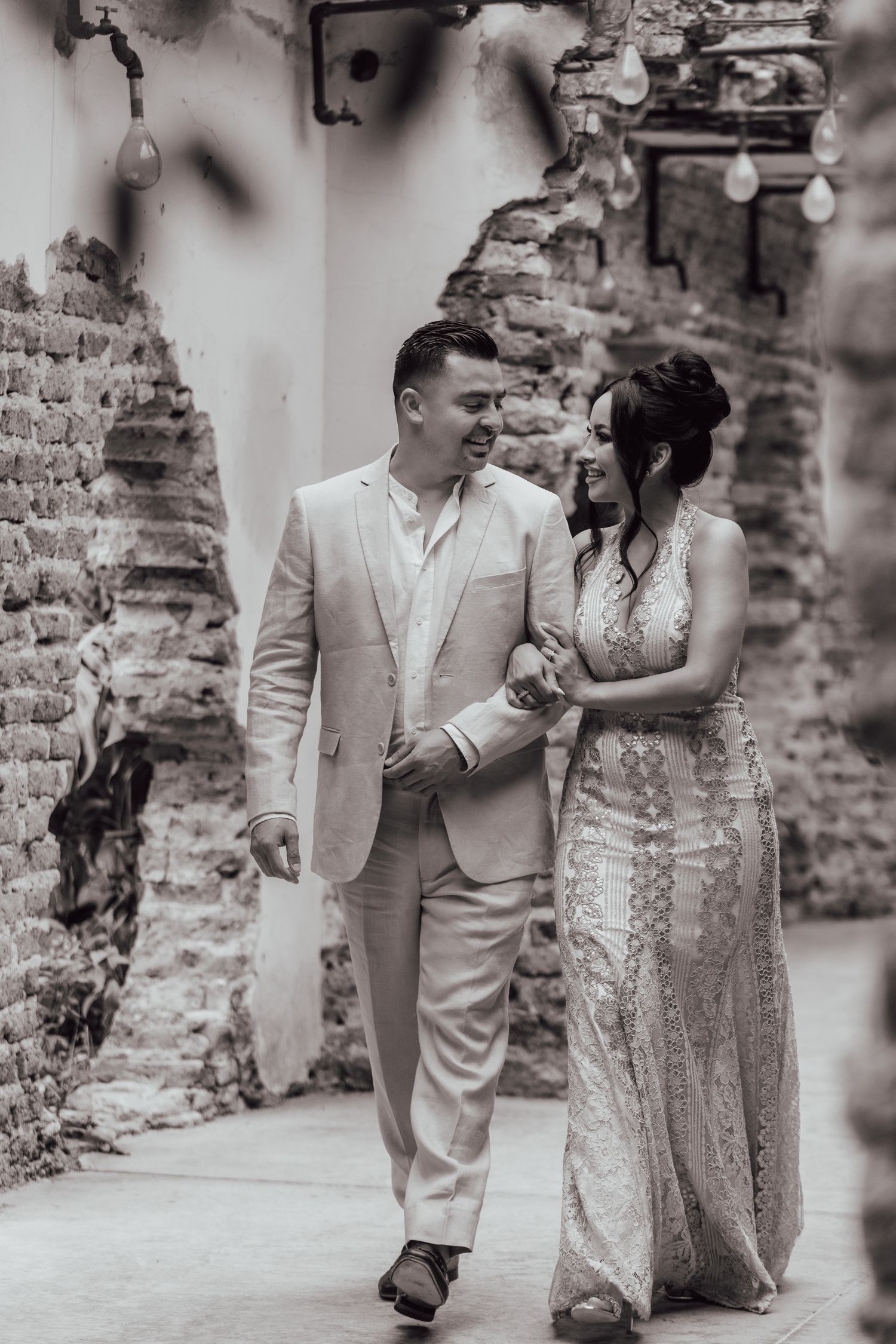 Black and white photo of a couple walking arm in arm through stone ruins, wearing elegant formal attire.