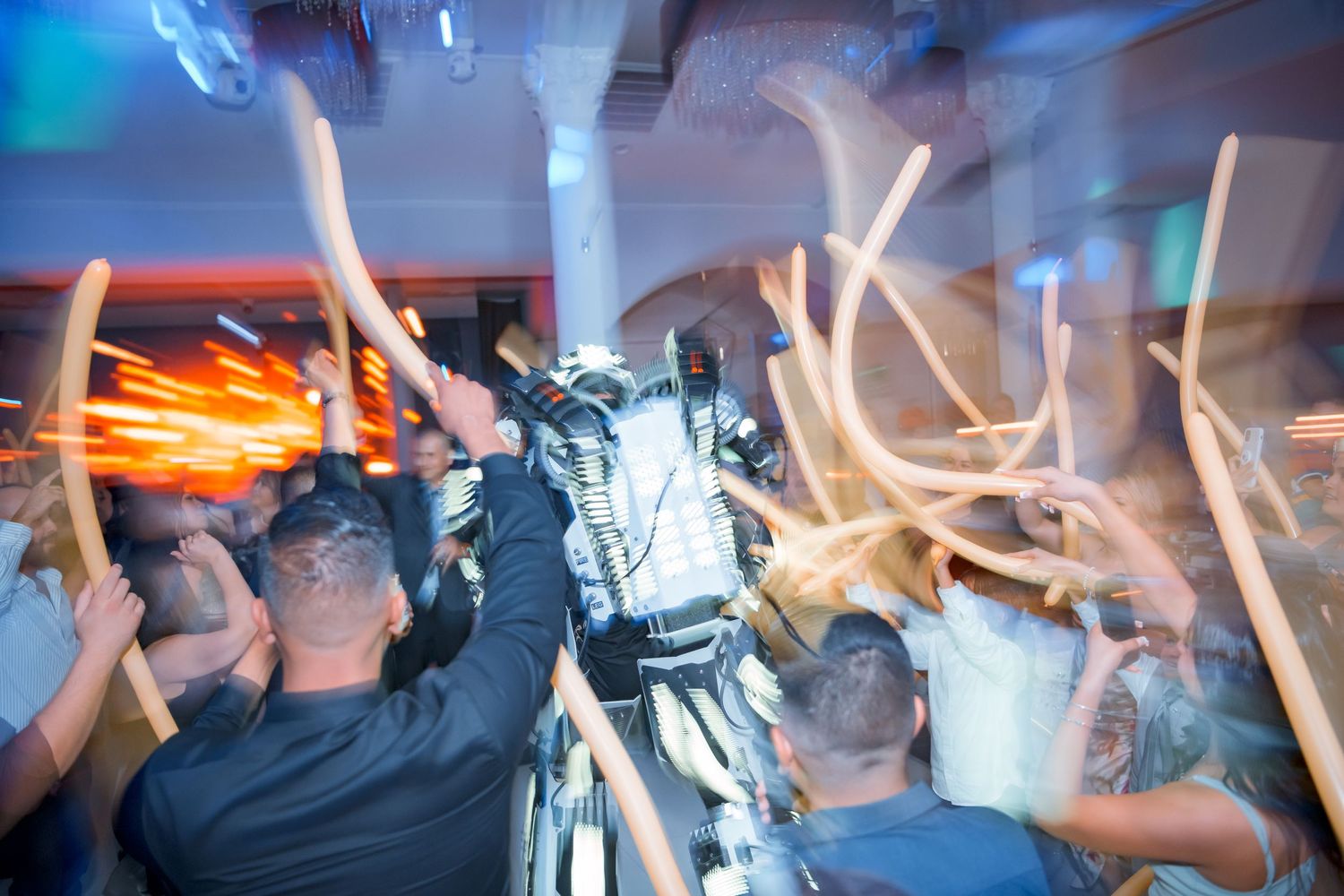 Motion blurred photo of a crowded dance floor with raised hands and glowing lights creating streaks in a nightclub atmosphere.