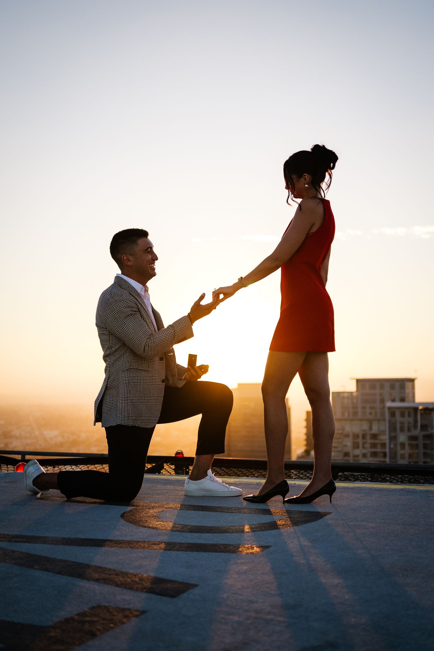 A silhouetted proposal scene at sunset on a rooftop with a figure kneeling and holding out a ring.