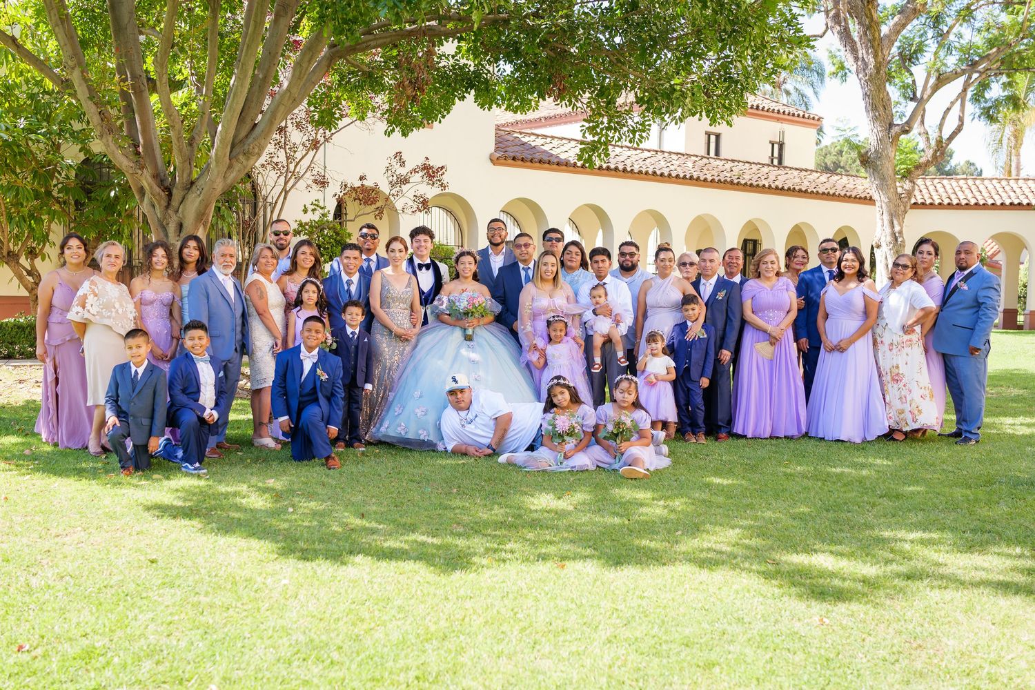 Large wedding party in blue and pink attire poses together on sunny lawn in front of Spanish-style building.