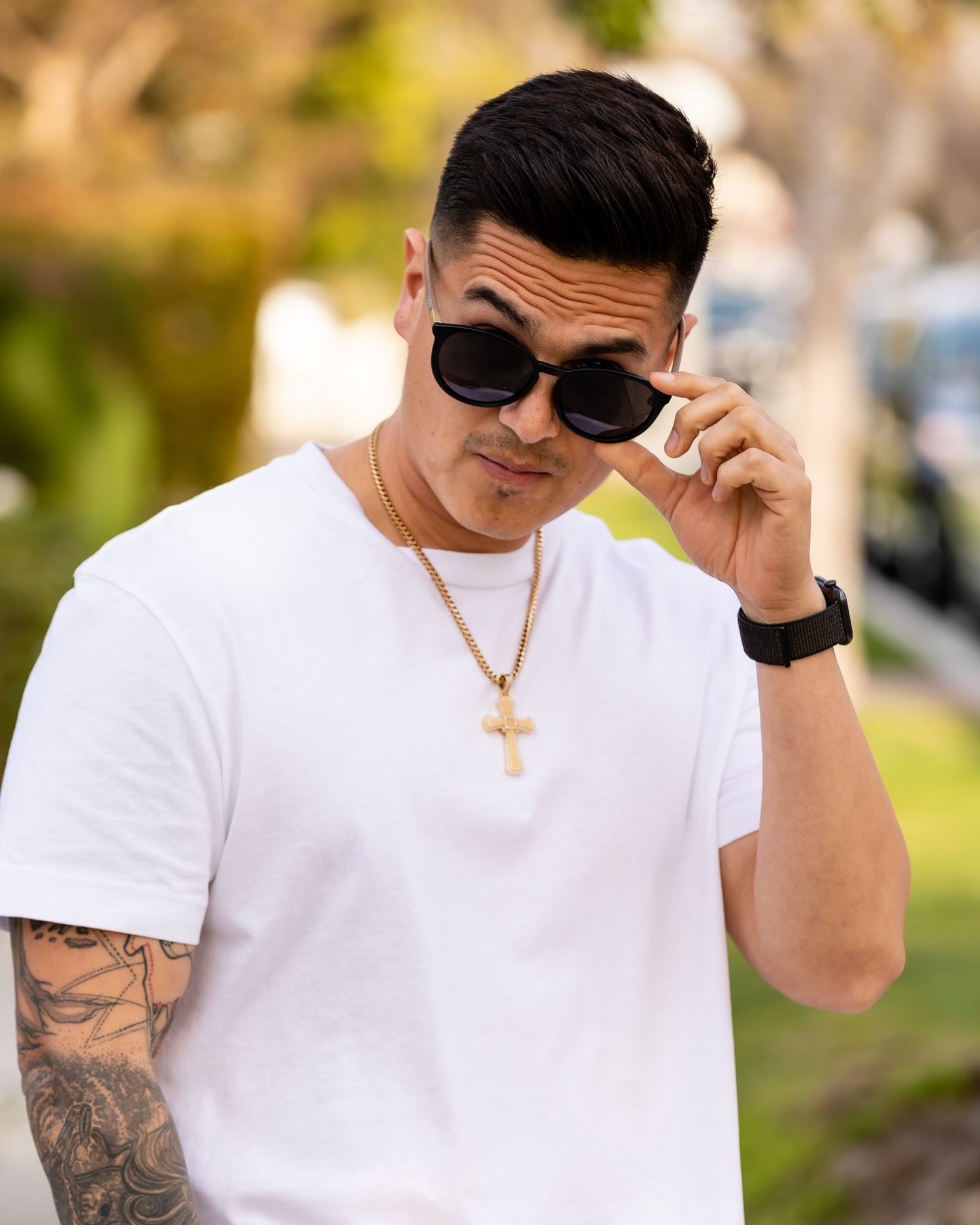 Man in white t-shirt and sunglasses with tattoos adjusting frames outdoors.