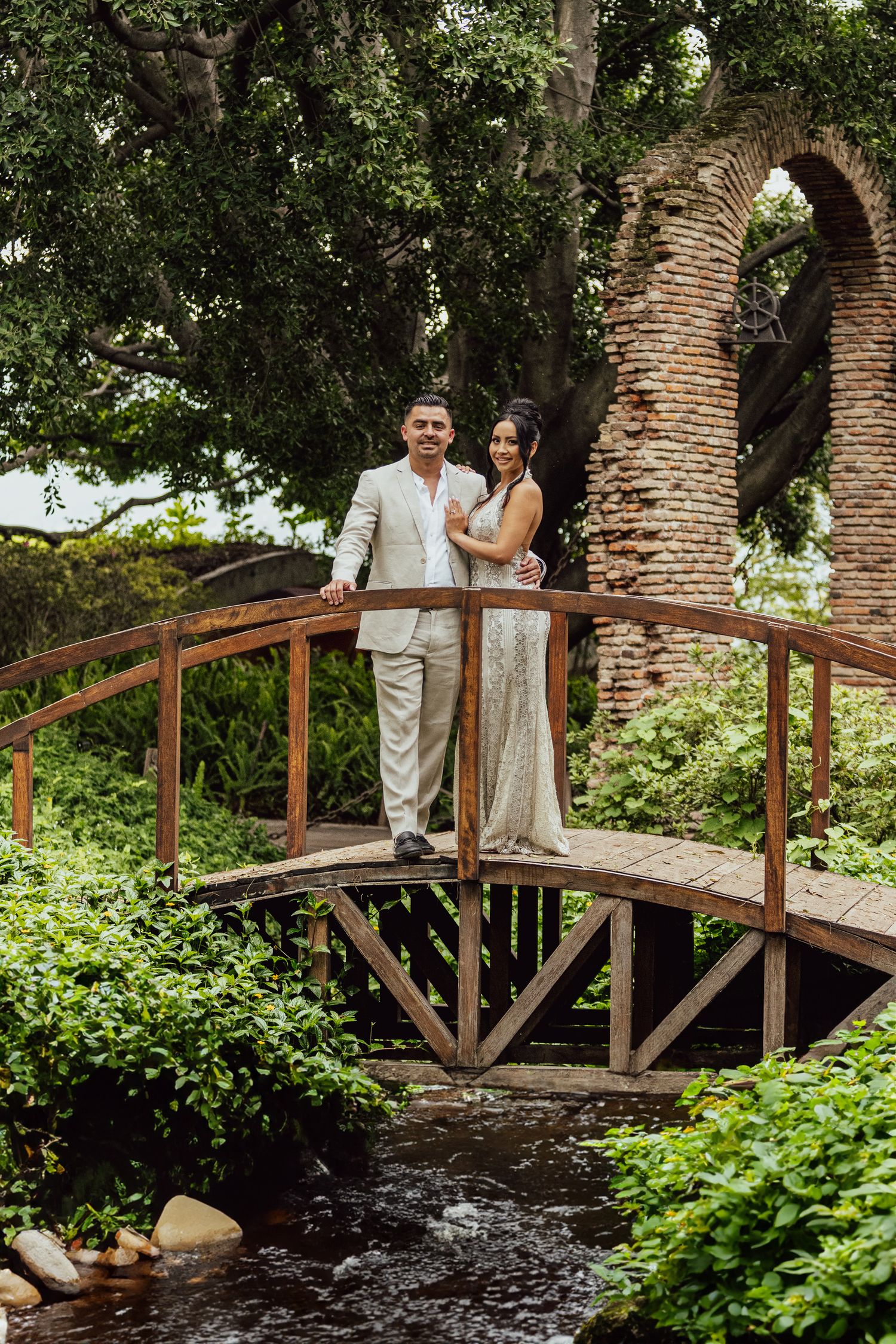 A couple stands on a rustic wooden bridge over a stream, surrounded by lush greenery and stone archways.