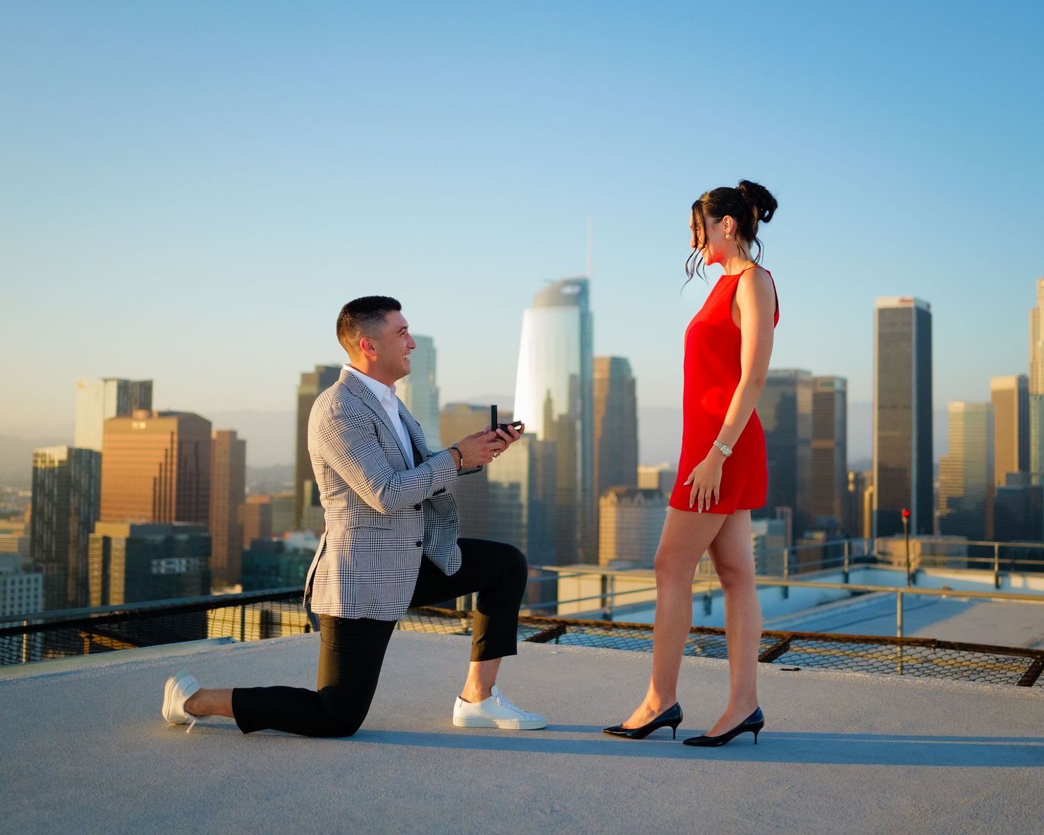 A romantic rooftop proposal scene at sunset with the downtown city skyline in the background.
